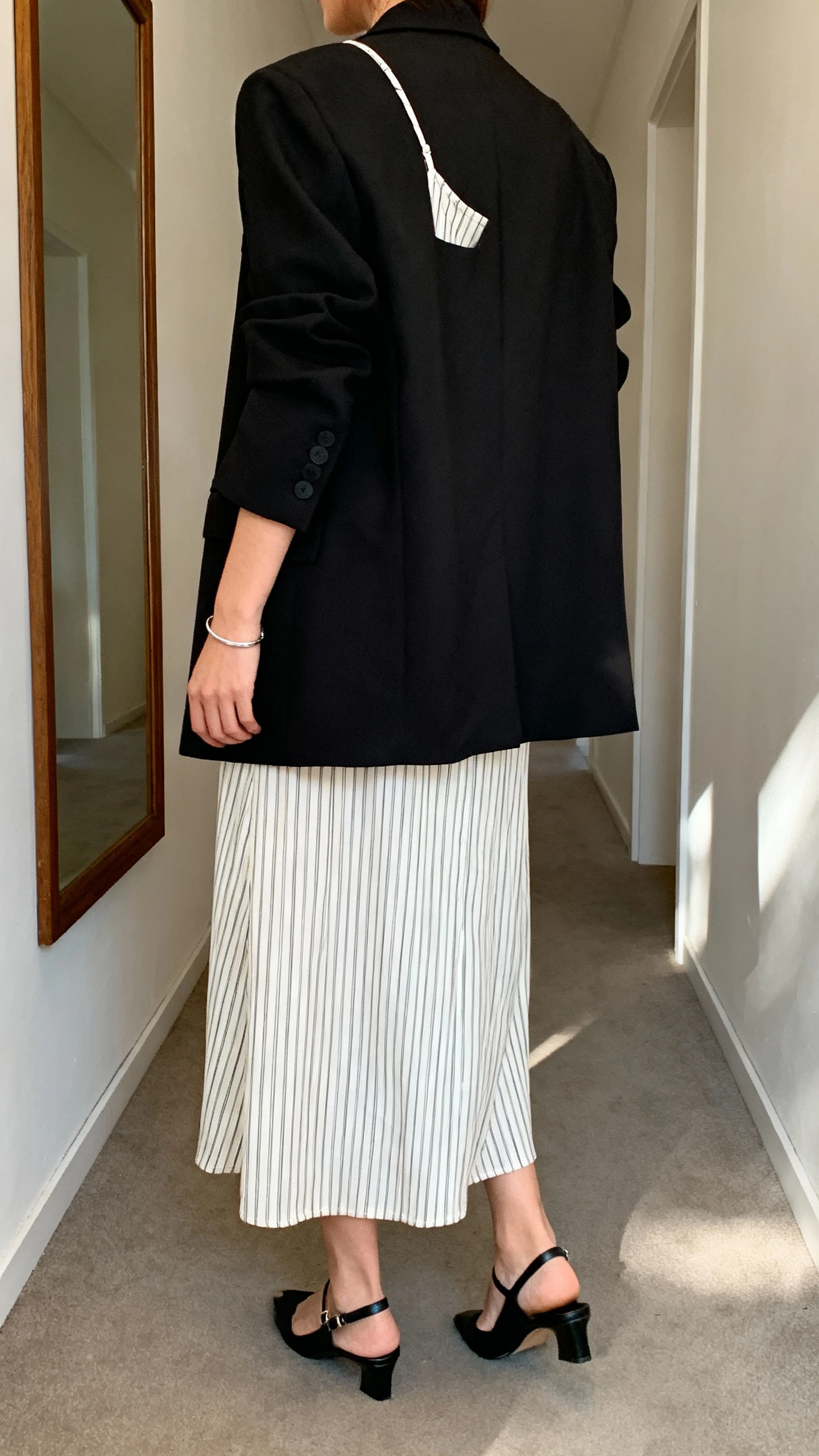 iPhone back view of a woman wearing a pinstriped midi slip dress layered under a black longline blazer, black slingback flats, delicate silver bracelet, face not visible, hallway mirror, natural daylight, iPhone photo quality.