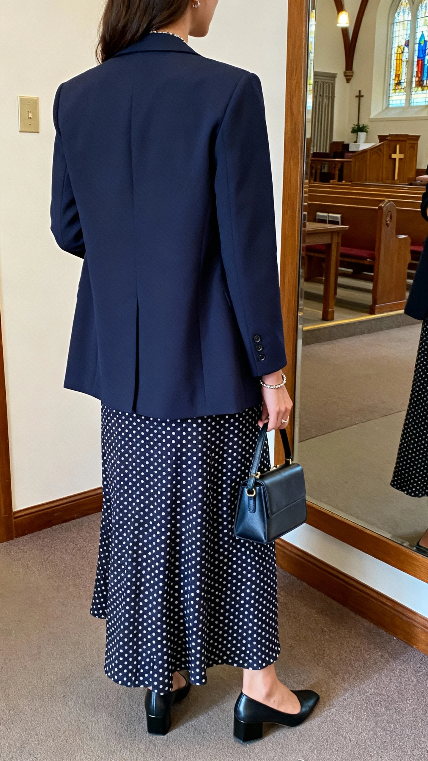 iPhone back view of a woman wearing a polka dot maxi dress with a tailored navy blazer, low block heels, small structured handbag, minimal jewelry, face not visible, church foyer mirror vibe with indoor lighting, iPhone photo quality
