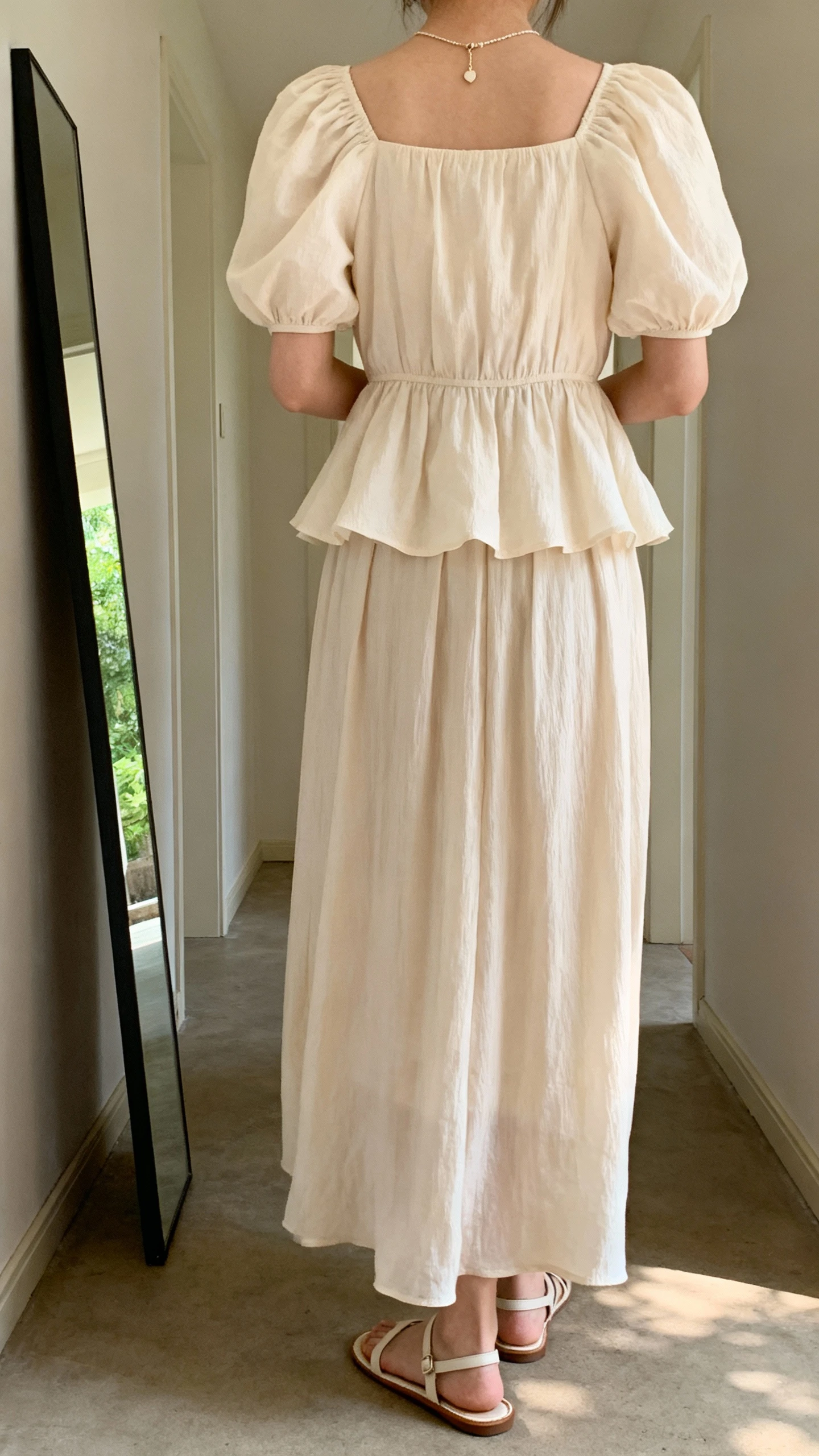 iPhone back view of a woman wearing a romantic puff-sleeve peplum blouse paired with a flowing maxi skirt, dainty pendant and simple sandals, face not visible, hallway full-length mirror, soft afternoon daylight, iPhone photo quality.