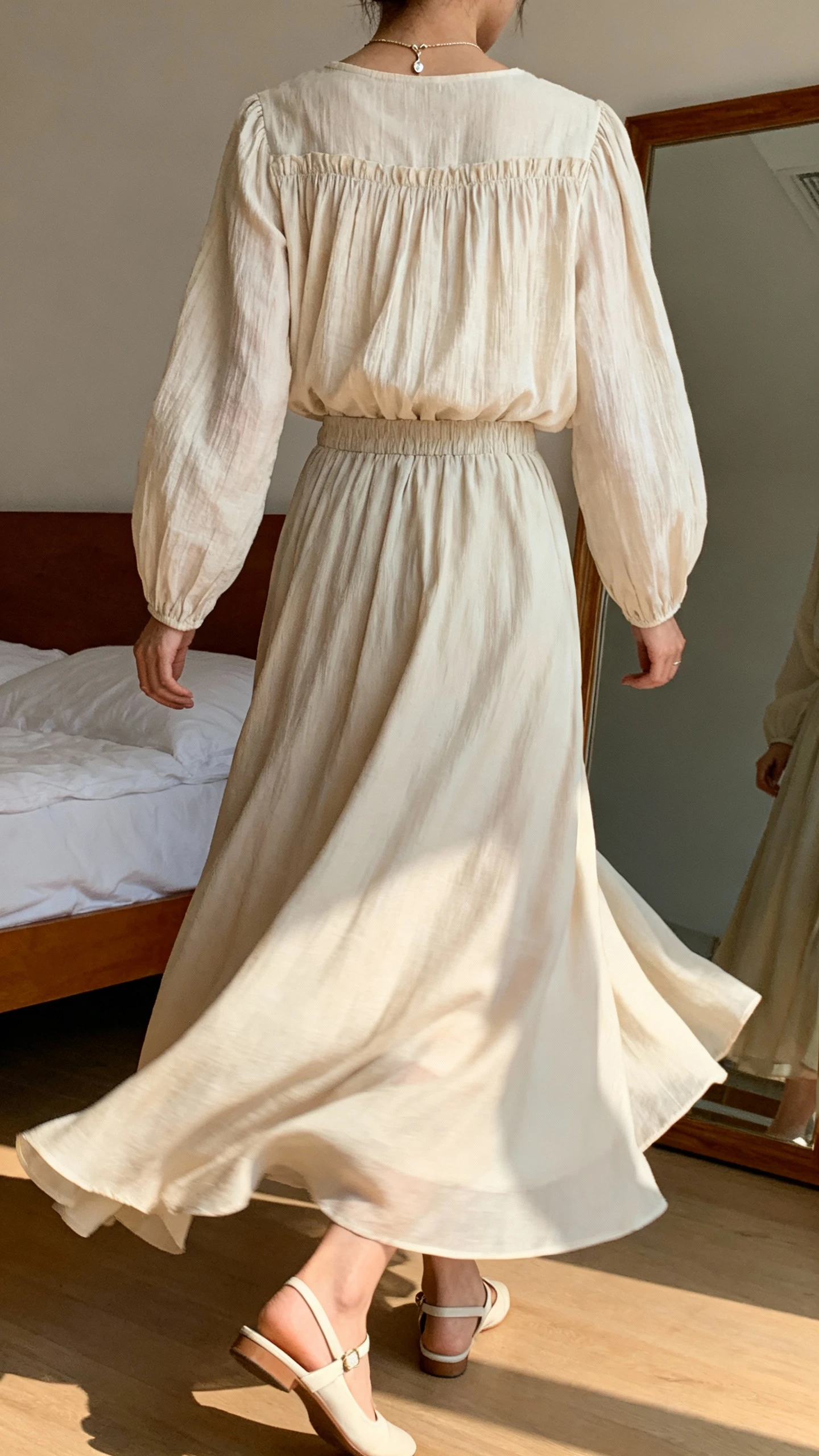 iPhone back view of a woman wearing a smocked long-sleeve blouse and a flowing maxi skirt that sways, simple ankle-strap flats and a dainty pendant, face not visible, walking pose by bedroom mirror, gentle afternoon light, iPhone photo quality.