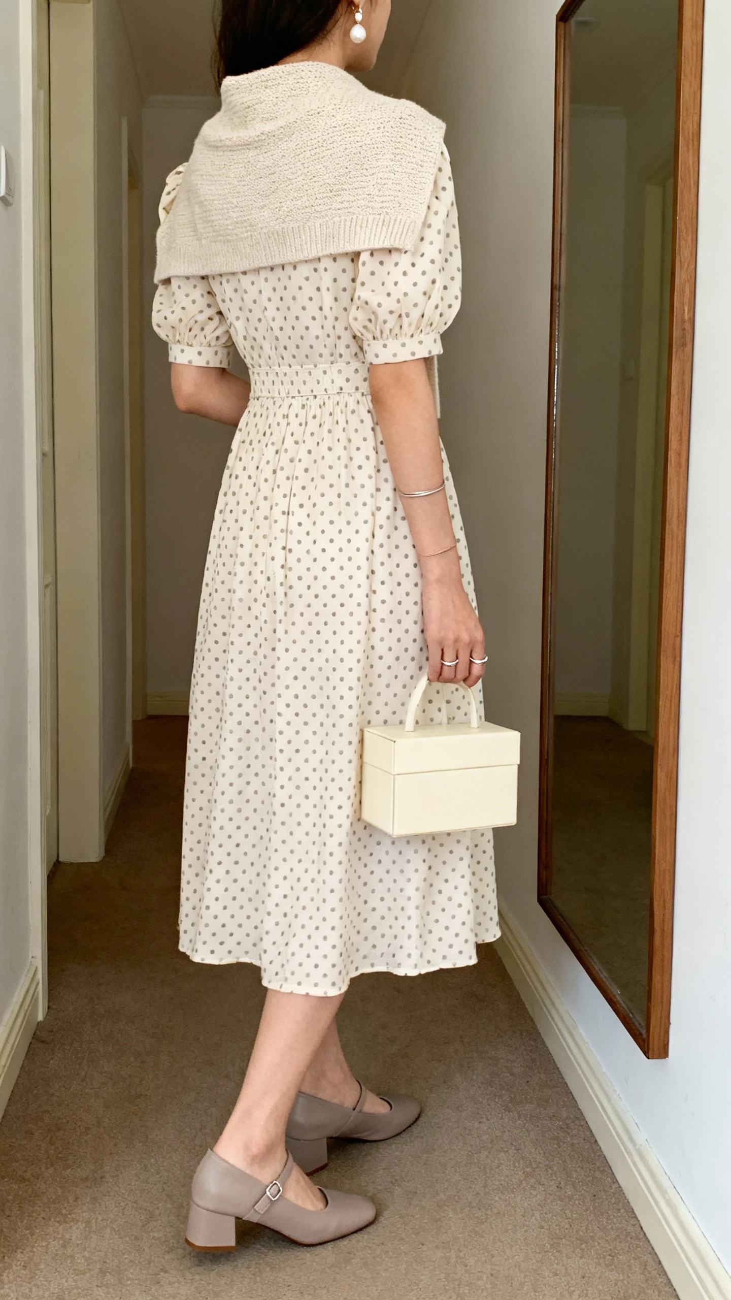 iPhone back view of a woman wearing a tea-length polka dot dress with modest puff sleeves and a gently defined waist, low-heel Mary Janes in soft taupe, a small cream box bag, pearl drop earrings, a thin stacking ring, and a fine-knit short cardigan over the shoulders, face not visible, hallway mirror, natural daylight, casual iPhone photo.