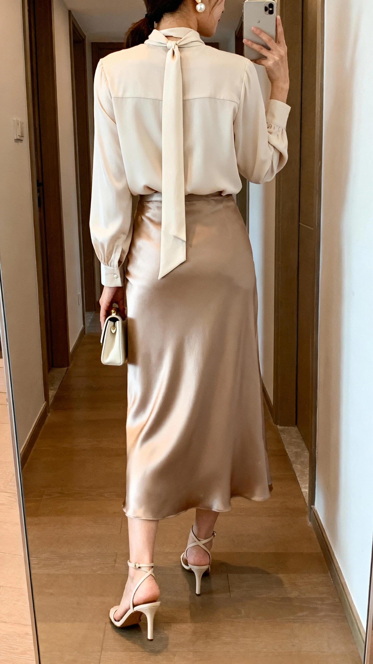 iPhone back view of a woman wearing a tie-neck blouse paired with a satin maxi skirt, slim ankle-strap heels, small clutch, pearl studs, face not visible, hallway mirror, warm indoor lighting, iPhone photo quality.