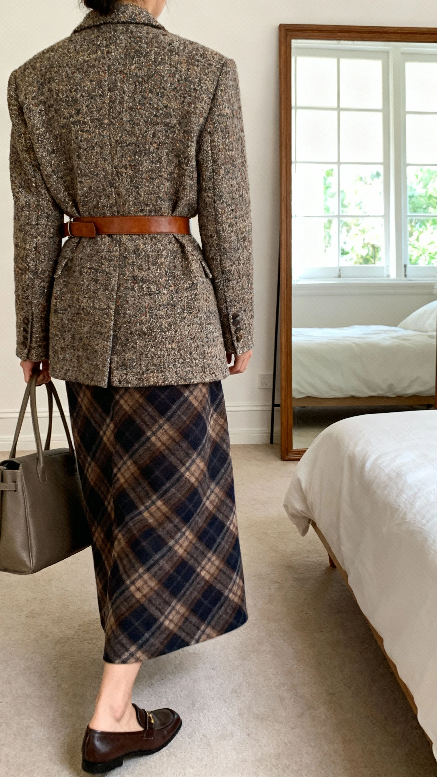 iPhone back view of a woman wearing a tweed blazer over a plaid maxi skirt, leather belt, loafers, and a structured tote, face not visible, walking pose toward a bedroom mirror, natural window light, iPhone photo quality.