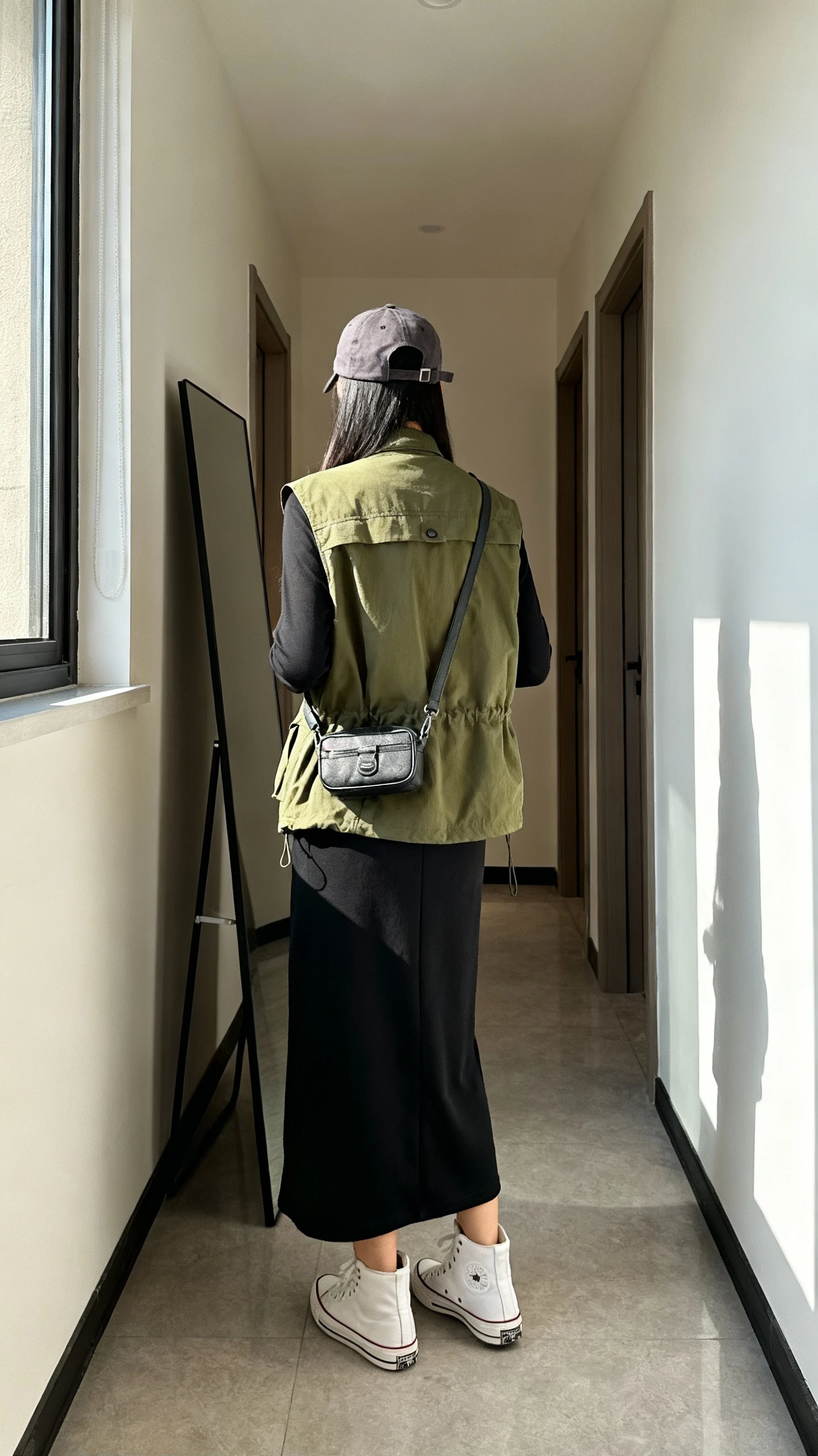 iPhone back view of a woman wearing an olive utility vest over a black jersey maxi dress with white high-top sneakers, baseball cap, crossbody belt bag, face not visible, hallway full-length mirror, natural daylight from window, iPhone photo quality.