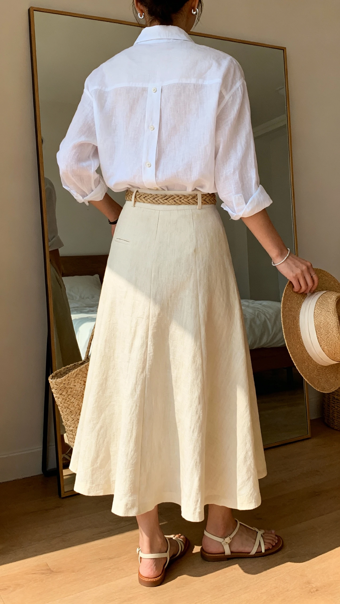 iPhone back-view photo of a woman in a cream full-length linen A-line skirt, white linen button-up shirt slightly tucked with a woven belt, strappy flat sandals, straw tote, lightweight stud earrings, dainty bracelet, and optional wide-brim hat in hand, face not visible, bedroom mirror, warm afternoon light, iPhone photo quality.
