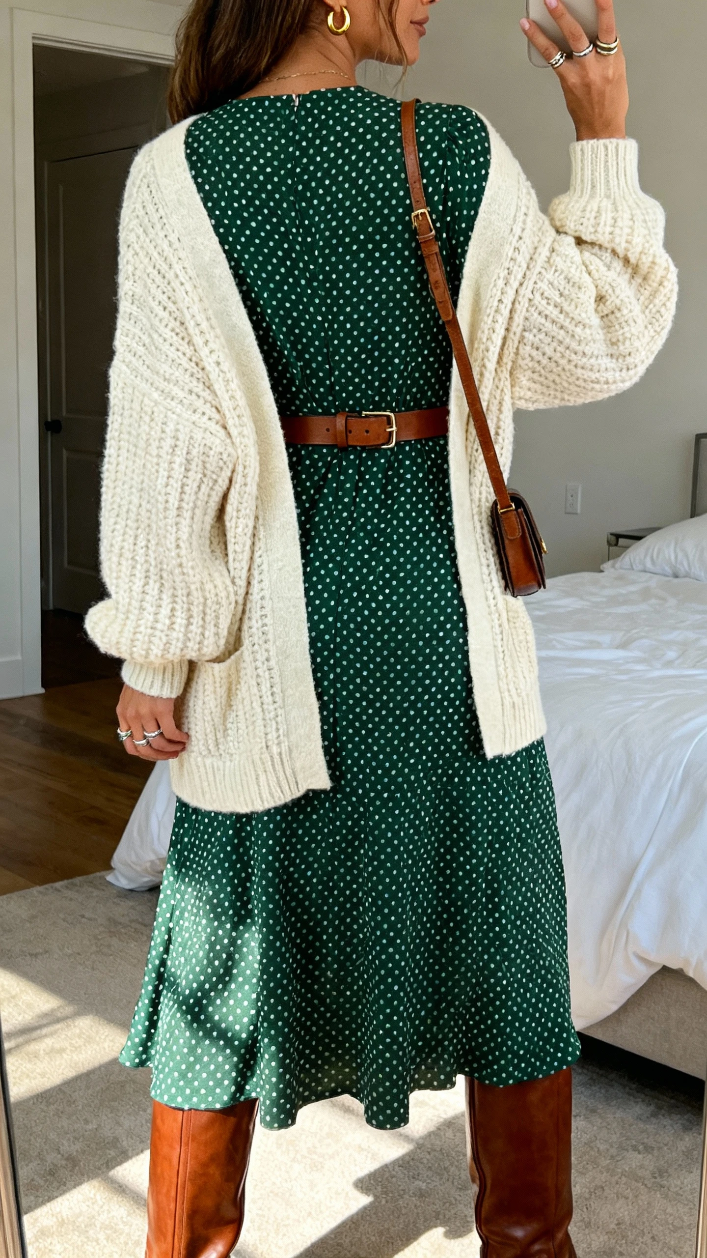 iPhone back-view photo of a woman in a forest green polka dot midi dress with a modest neckline, thick cream knit cardigan belted at the waist with a leather belt, knee-high brown leather boots, gold huggie earrings, stackable rings, and a small crossbody, face not visible, bedroom mirror, soft afternoon light, iPhone photo quality.