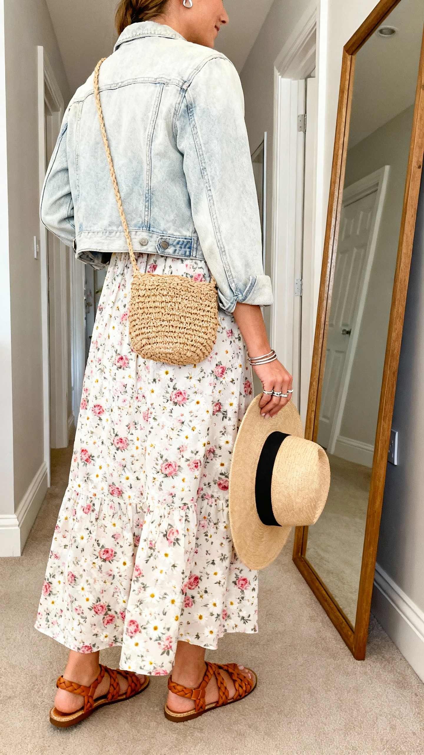 iPhone back-view photo of a woman wearing a light floral maxi dress (peony or tiny daisy print) with a soft light-wash denim jacket, braided flat sandals (tan or caramel), a woven straw or raffia crossbody bag, stackable rings and simple stud earrings, and an optional wide-brim hat in hand, face not visible, hallway full-length mirror, natural daylight, iPhone photo quality.