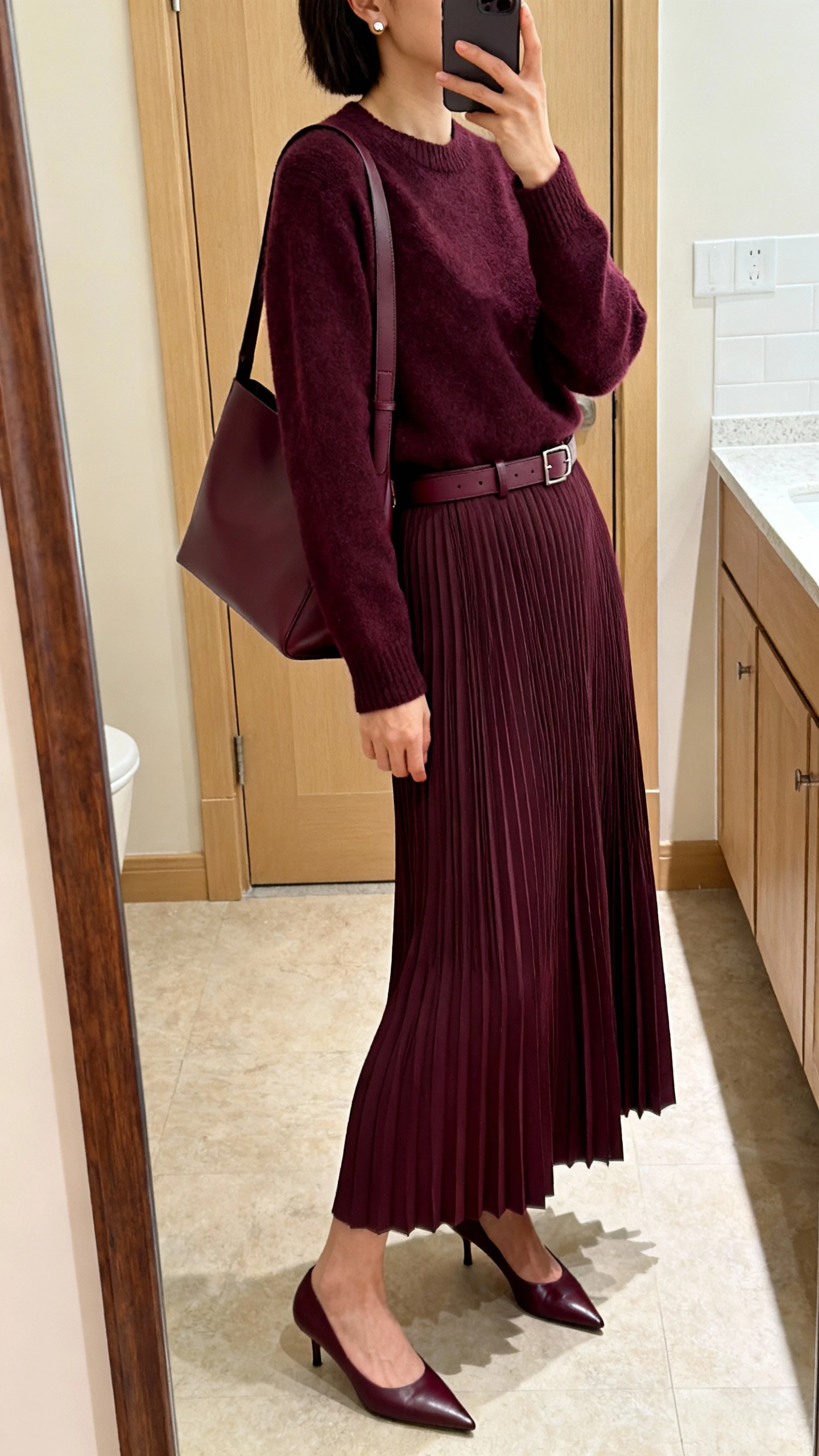 iPhone bathroom mirror selfie of a woman wearing a fine-knit crewneck in deep plum with a matching tonal pleated maxi dress (micro pleats), a minimal leather belt in the same color family, pointed kitten heels, simple stud earrings, and a medium shoulder bag with clean lines; face not visible, side angle, indoor lighting, iPhone photo quality.