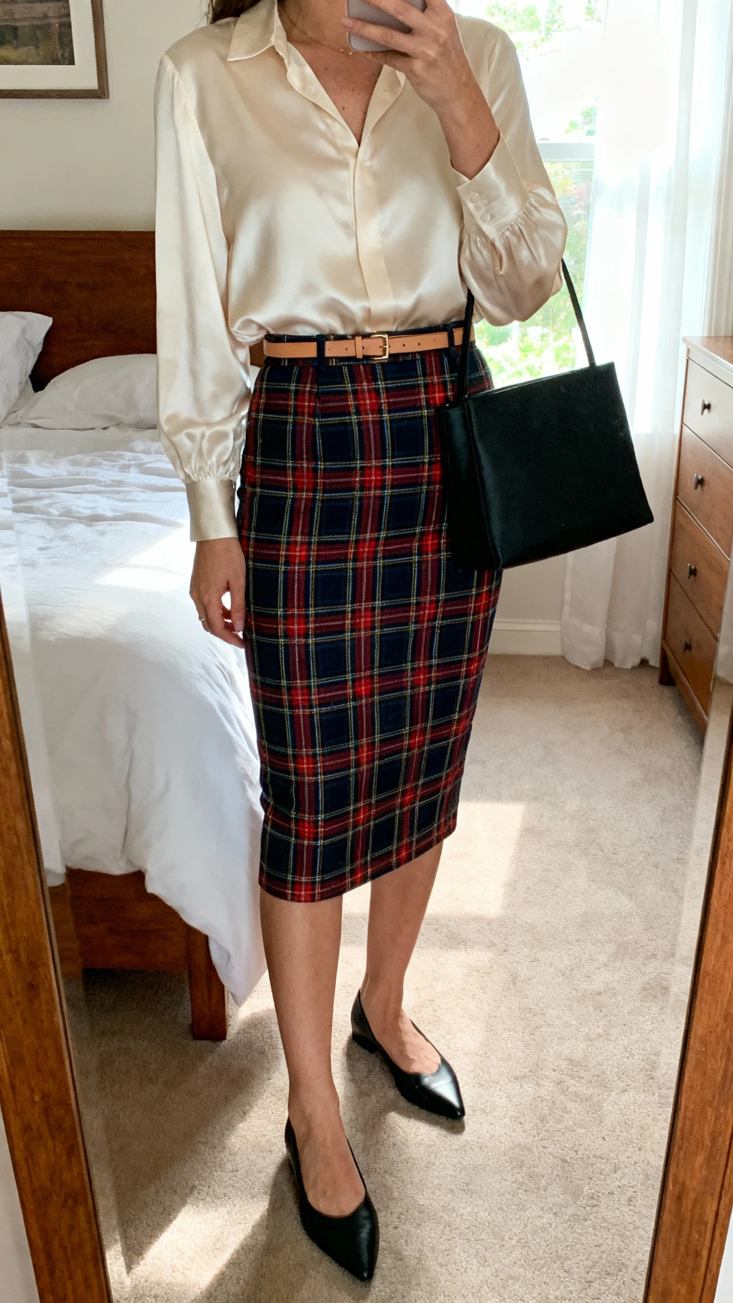 iPhone mirror selfie of a woman in a below-the-knee tartan pencil skirt, ivory satin-look blouse, optional thin leather belt matching black pointed-toe flats, and a minimal black tote; face not visible, cropped at shoulders, bedroom mirror, soft afternoon window light, iPhone photo quality.