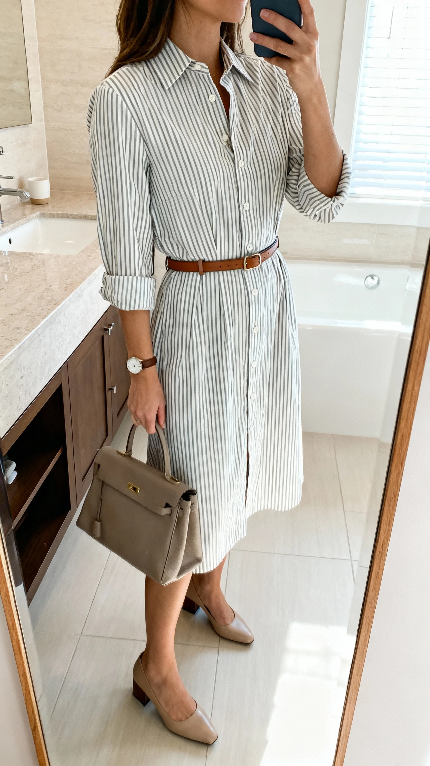iPhone mirror selfie of a woman in a belted striped shirt dress (button-front, cuffed sleeves, crisp cotton, knee-to-midi length) with a slim leather belt, low block-heel pumps, structured tote, and a simple watch; face not visible, side angle, bathroom mirror, bright indoor lighting, iPhone photo quality.