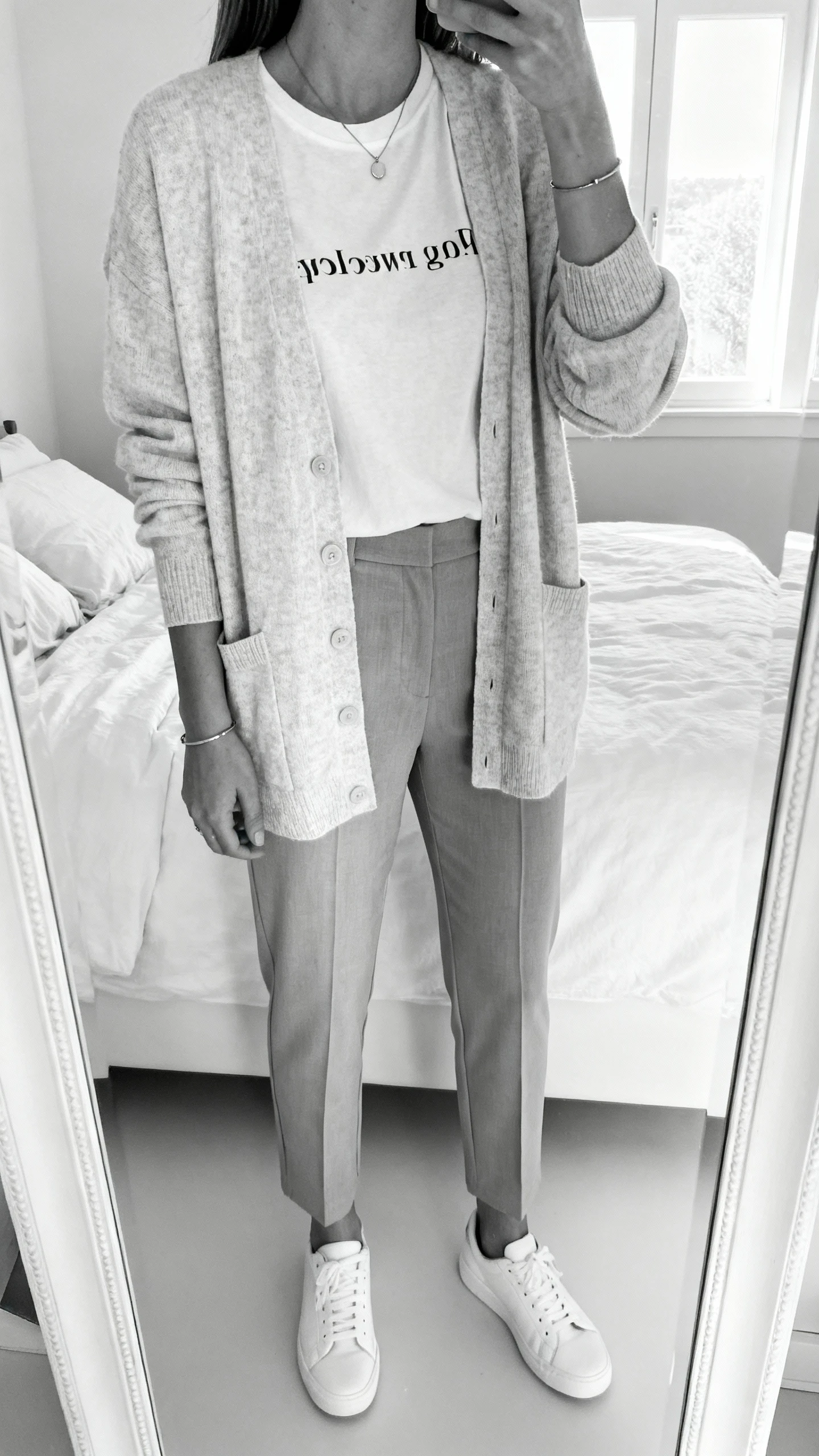 iPhone mirror selfie of a woman in a clean monochrome set: soft cardigan over a subtle message tee, matching tailored pants, minimalist white sneakers, slim pendant necklace, and simple bracelet; face not visible, cropped at neck, bedroom mirror, bright natural window light, iPhone photo quality.