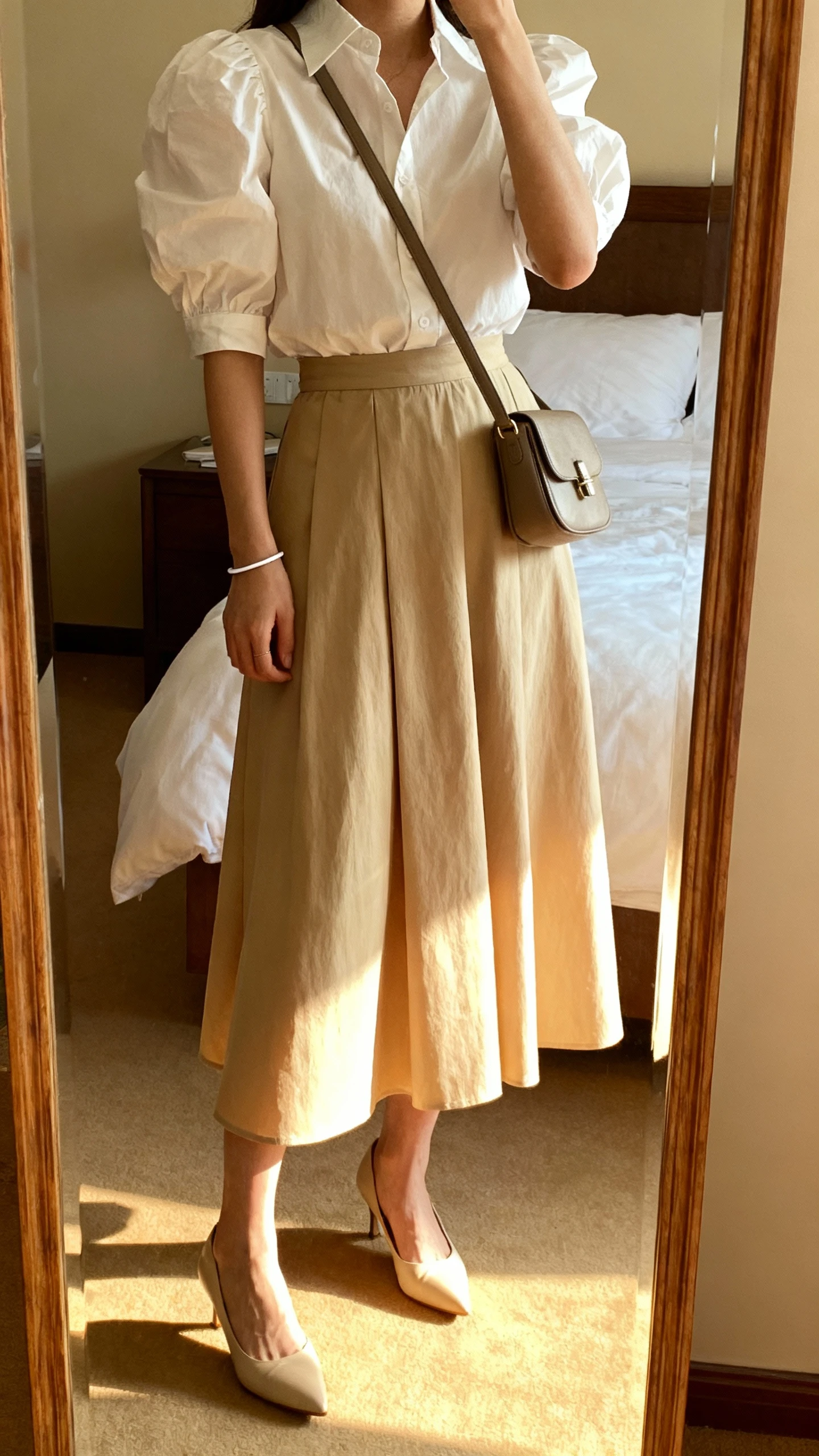 iPhone mirror selfie of a woman in a crisp poplin puff-sleeve blouse tucked into a tea-length skirt, kitten heels, structured crossbody, and a thin bracelet, face not visible, cropped at shoulders, bedroom mirror, soft afternoon light, iPhone photo quality.
