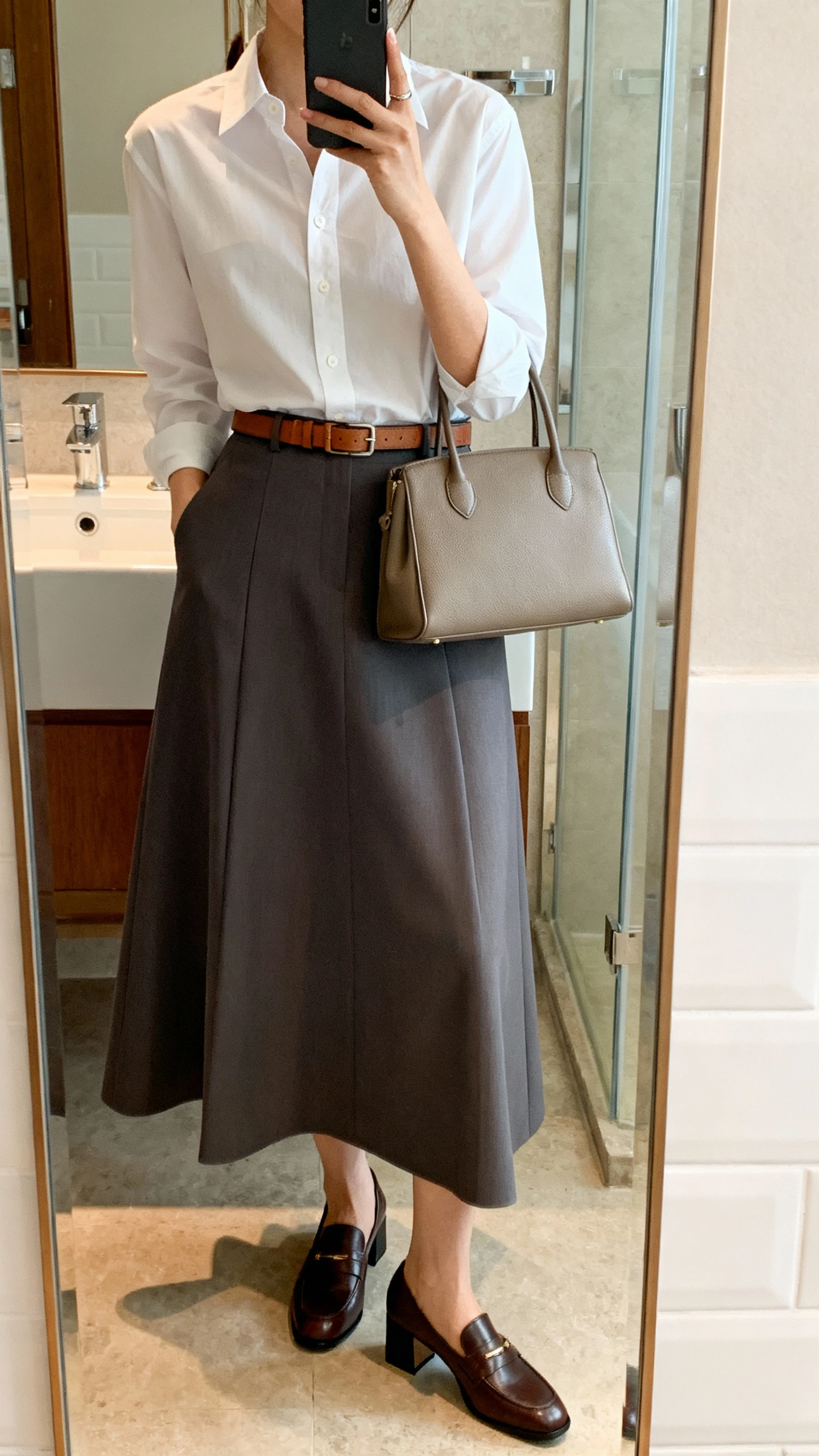 iPhone mirror selfie of a woman in a crisp white button-down tucked into an A-line maxi skirt, slim leather belt, structured tote, and block-heel loafers, face not visible, bathroom full-length mirror, indoor lighting, iPhone photo quality.
