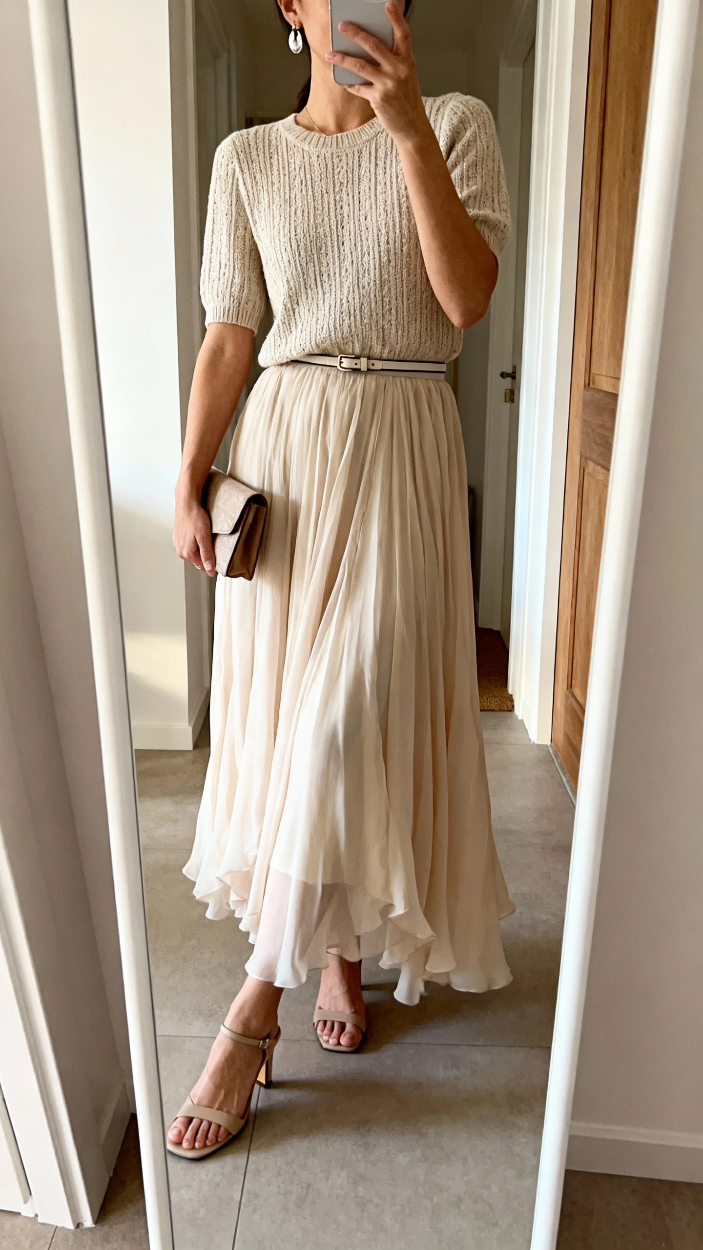 iPhone mirror selfie of a woman in a flowy chiffon maxi skirt with a structured knit top, heeled closed-toe sandals, minimal drop earrings, a compact clutch, and a thin belt at the waist; face not visible, cropped at neck, entryway mirror, soft indoor daylight, iPhone photo quality.