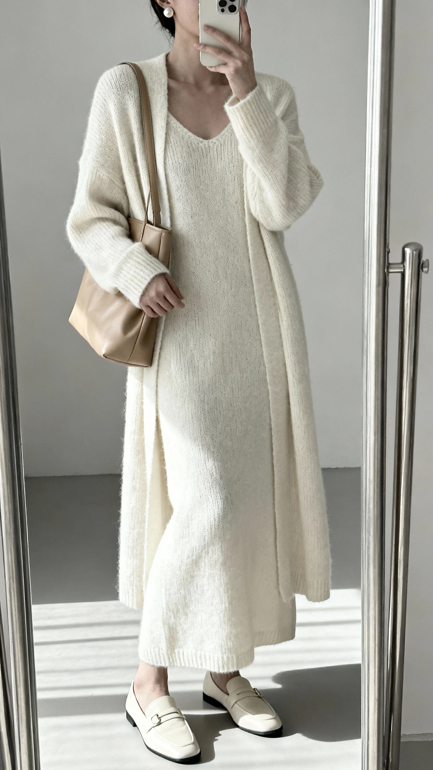 iPhone mirror selfie of a woman in a minimalist monochrome look: cream knit maxi dress, matching cream longline cardigan, beige leather shoulder bag, cream loafers, and subtle pearl studs, face not visible, cropped at neck, full-length mirror, gentle morning light, iPhone photo quality.