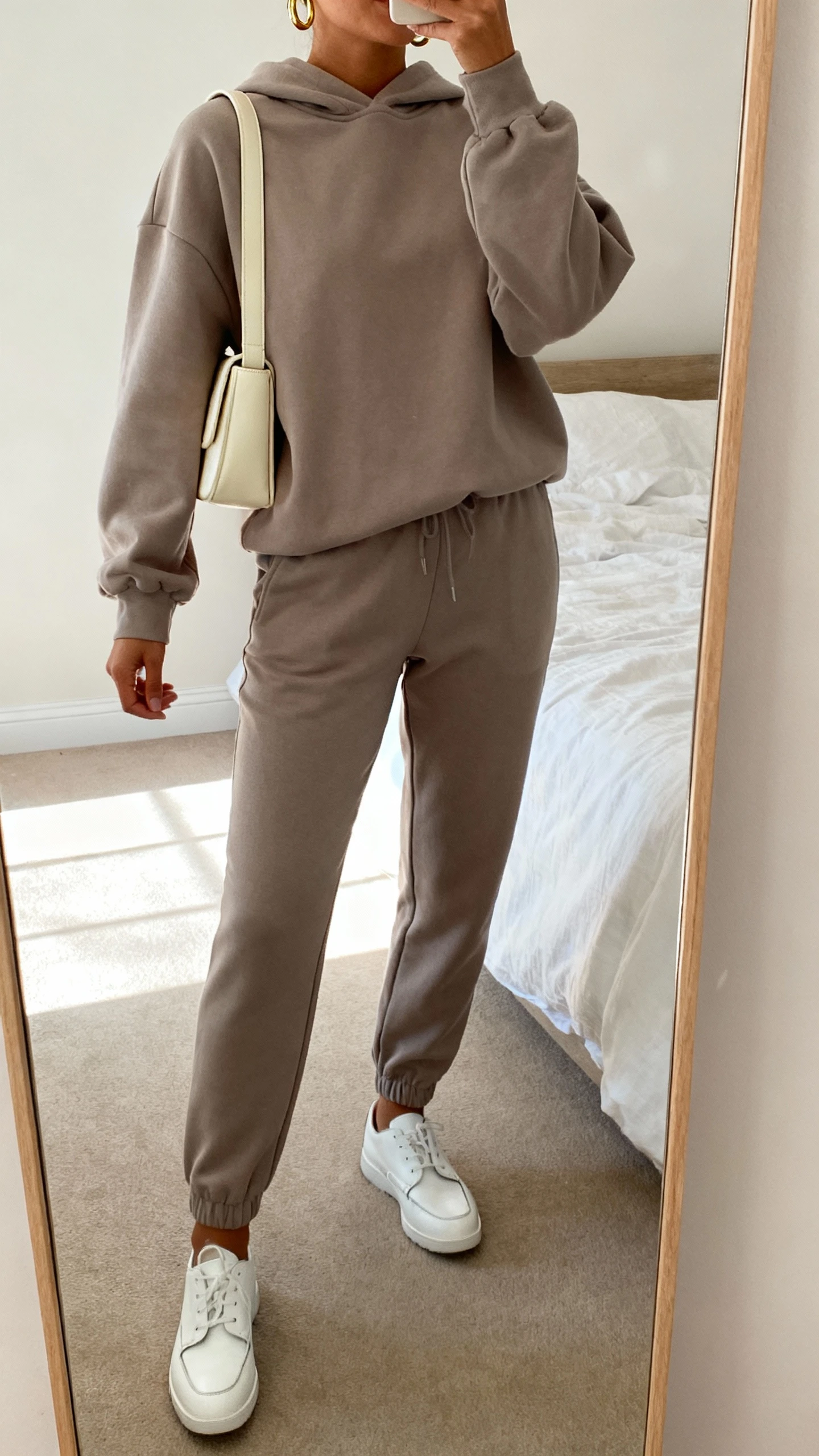 iPhone mirror selfie of a woman in a monochrome taupe tracksuit (relaxed hoodie and tapered joggers), sleek white leather trainers, minimal gold hoops, and a structured cream shoulder bag; face not visible, cropped at shoulders, bedroom mirror, soft afternoon light, iPhone photo quality.