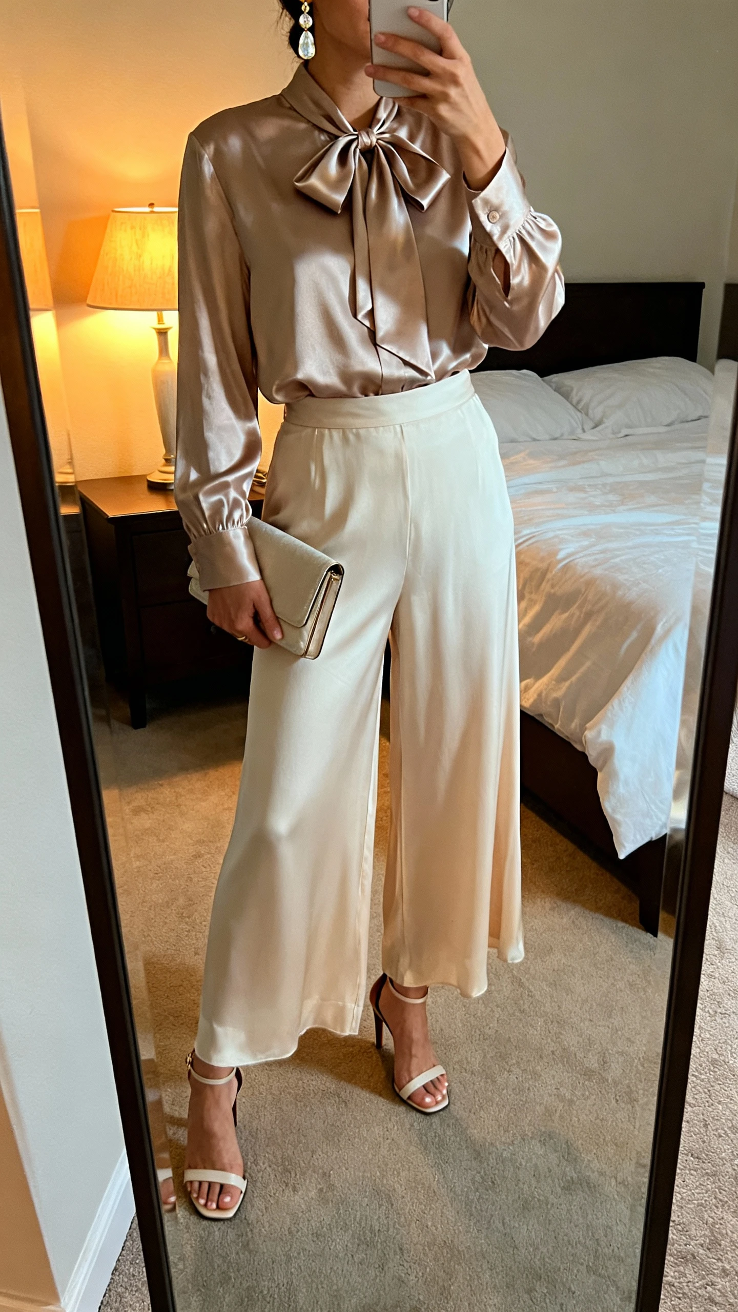 iPhone mirror selfie of a woman in a satin bow-tie blouse and flowing palazzo pants, heeled sandals, drop earrings, and a clutch, face not visible, cropped at shoulders, bedroom mirror with warm evening lamp light, iPhone photo quality.