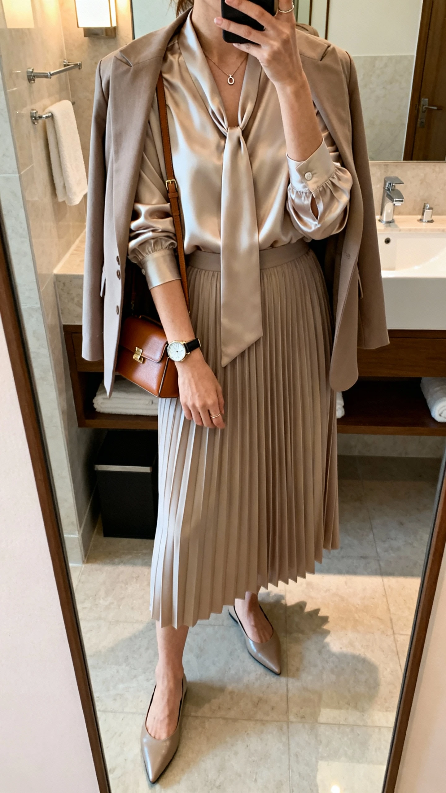 iPhone mirror selfie of a woman in a silky tie-neck blouse with sleeves tucked into a pleated midi skirt, pointed-toe flats, a minimal pendant necklace, a watch, a compact shoulder bag, and a lightweight blazer over her shoulders, face not visible, bathroom full-length mirror, soft indoor lighting, iPhone photo quality.