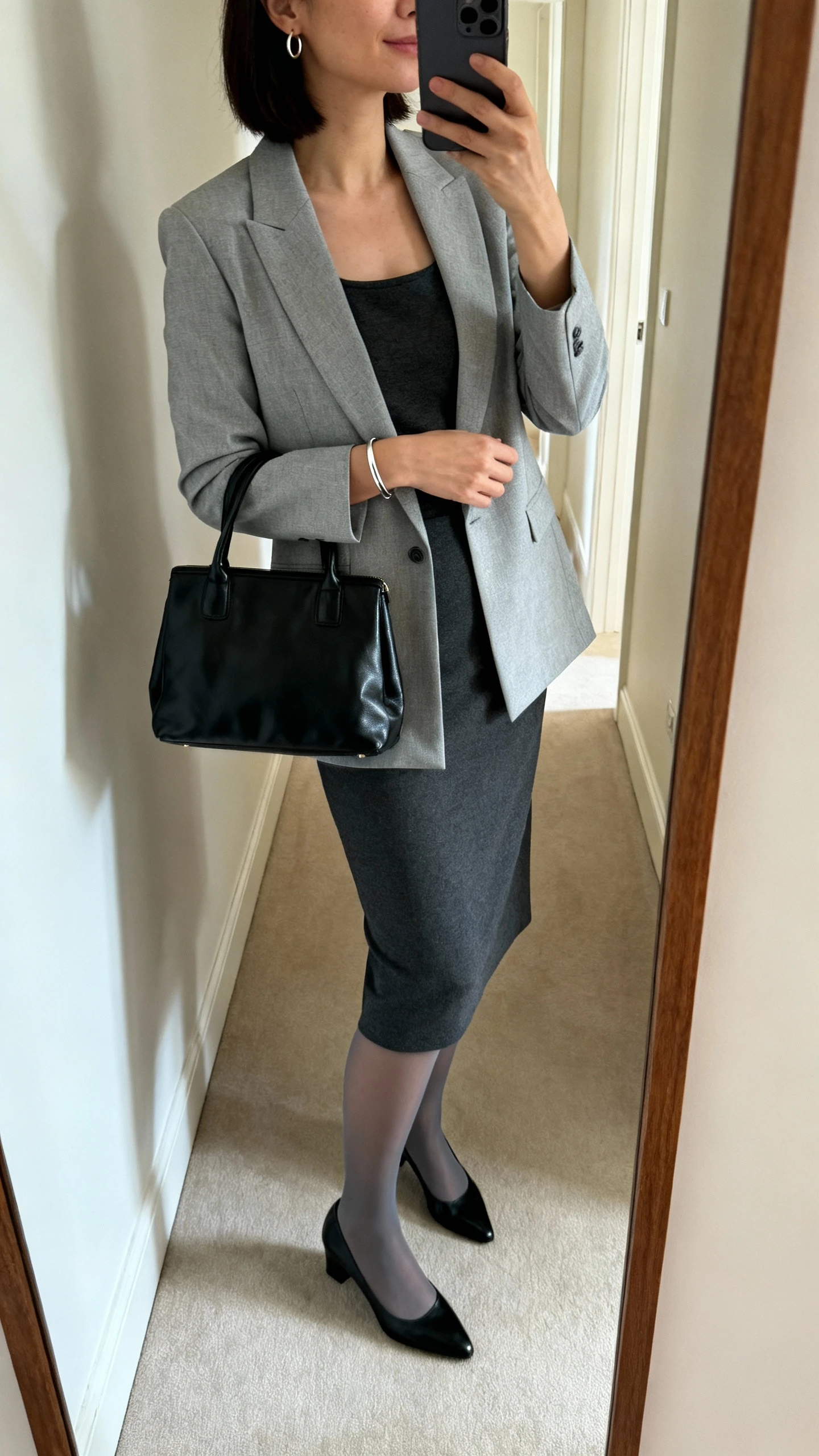iPhone mirror selfie of a woman in a solid charcoal sheath dress with a modest boat neckline, a light blazer in dove gray, classic black low-heel pumps, a structured black leather satchel, a thin silver bracelet with small hoop earrings, and optional sheer graphite tights; face not visible, side angle in hallway mirror, morning indoor light, iPhone photo quality.