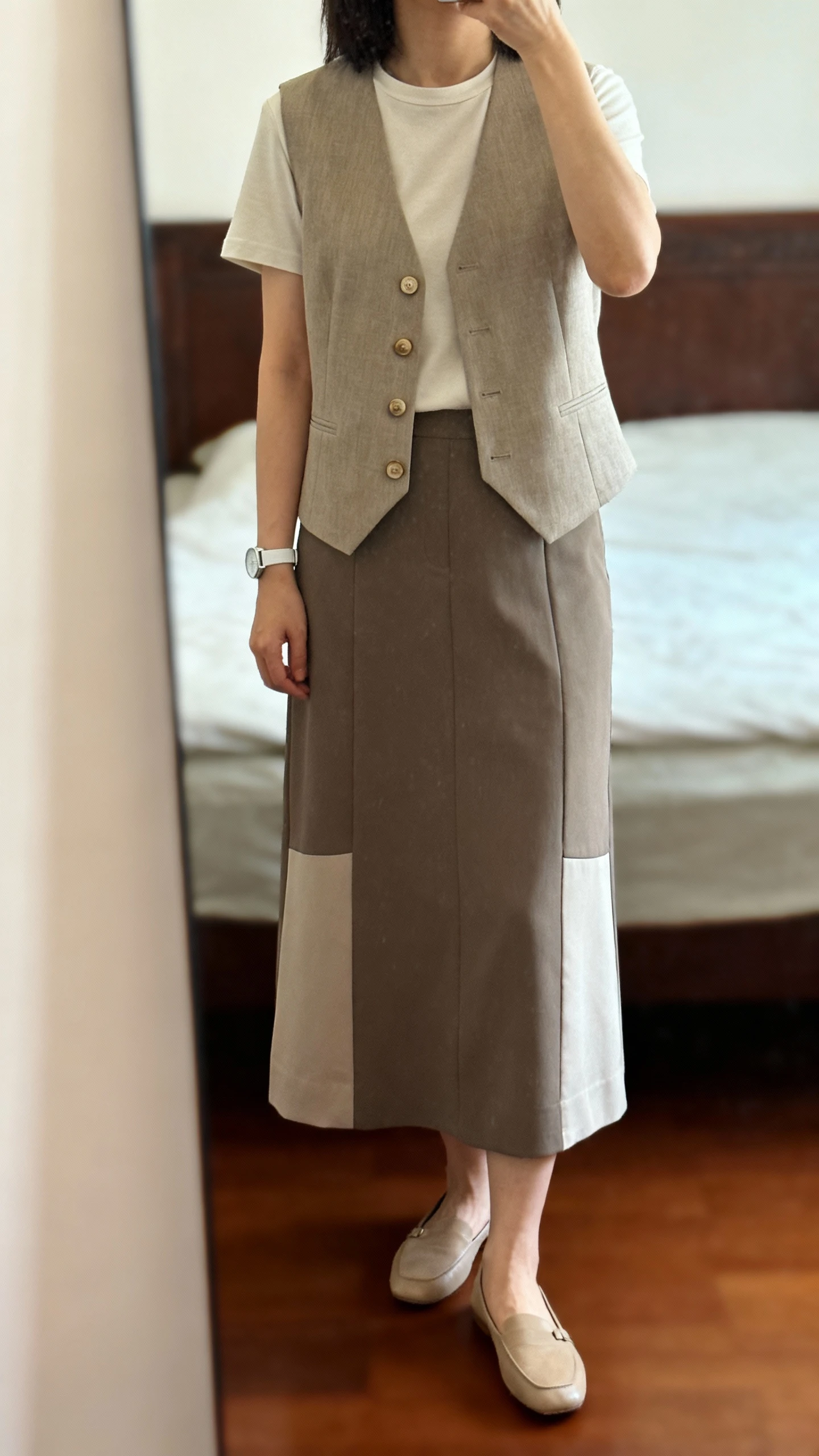 iPhone mirror selfie of a woman in a tailored buttoned vest-blouse layered over a soft tee and paired with a paneled midi skirt, comfortable flats, and a simple watch, face not visible, cropped at shoulders, bedroom mirror, warm indoor lighting, iPhone photo quality.