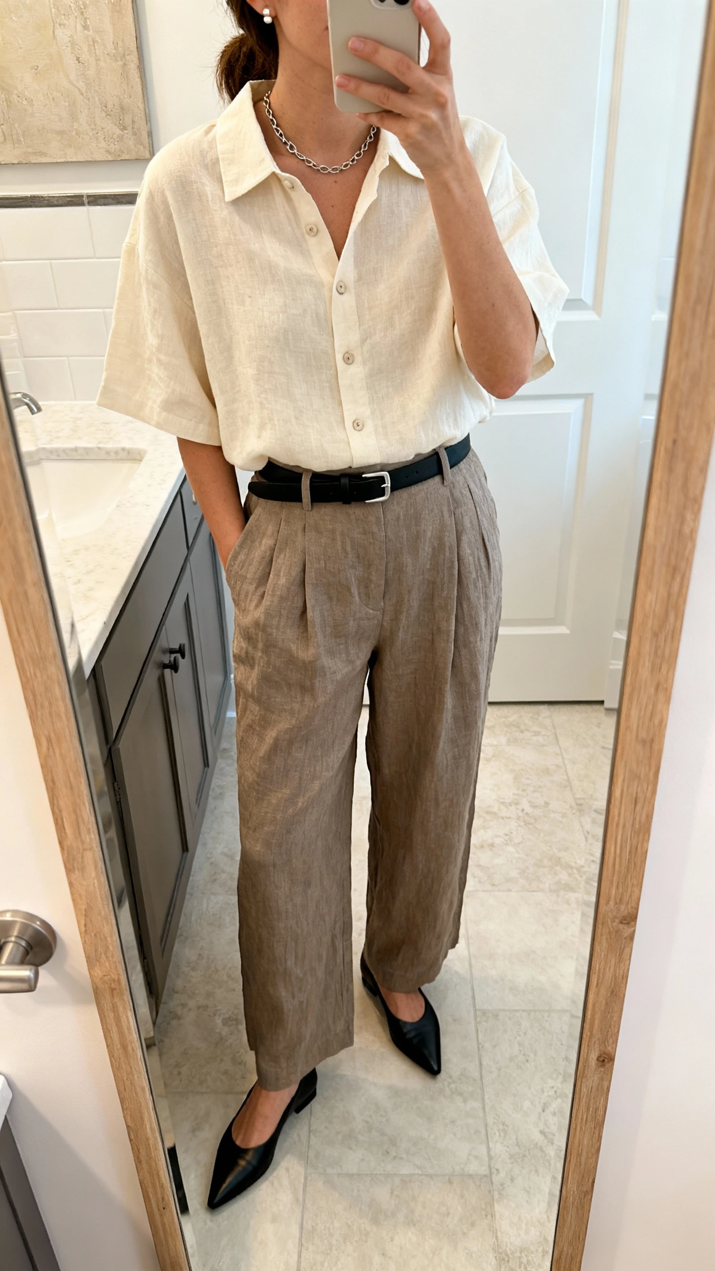 iPhone mirror selfie of a woman in high-waisted linen wide-leg trousers in taupe, a boxy short-sleeve linen button-up in cream, sleek pointed-toe flats in black, a minimal leather belt in black with a simple buckle, and a thin chain necklace with small stud earrings, face not visible, bathroom mirror, soft indoor lighting, iPhone photo quality.