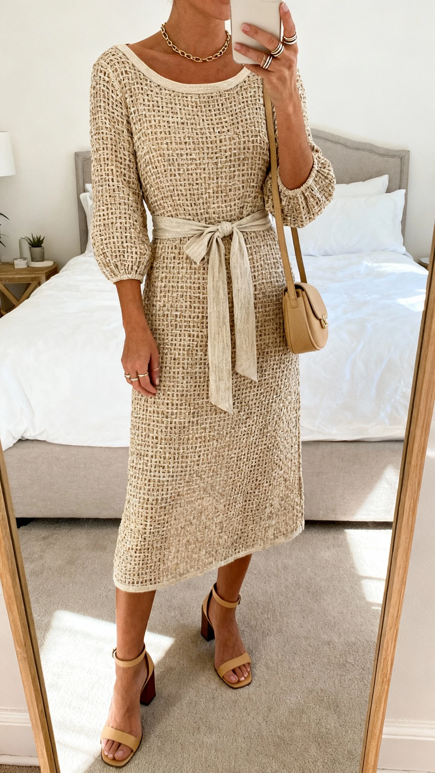 iPhone mirror selfie of a woman wearing a basket-weave midi dress with elbow sleeves and a gentle boatneck, tied with a fabric sash, ankle-strap block heels, a slim chain necklace, stackable rings, and a small shoulder bag in sand; face not visible, cropped at shoulders, bedroom mirror, natural daylight, iPhone photo quality.