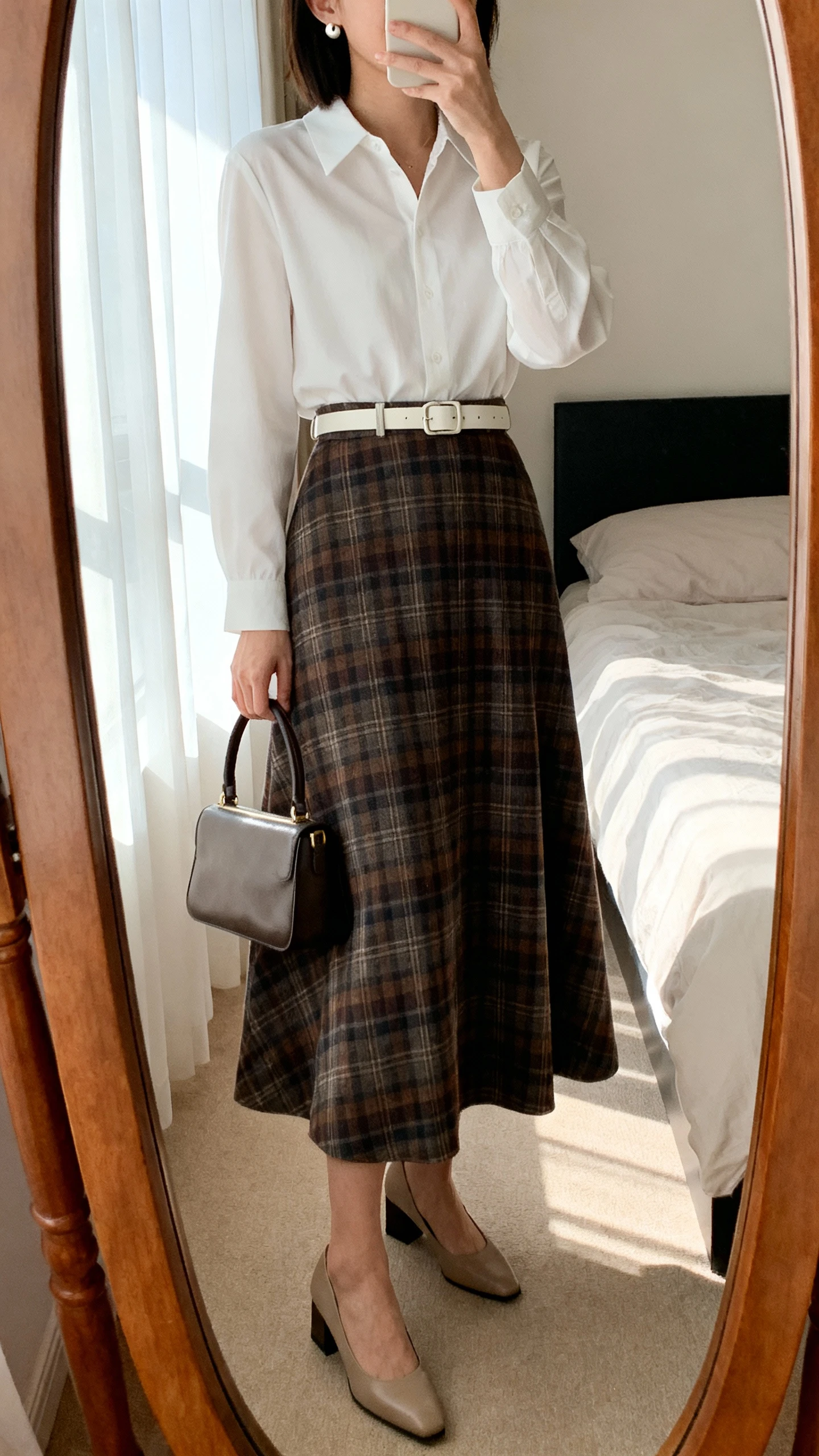 iPhone mirror selfie of a woman wearing a belted white blouse tucked into a classic A-line plaid long skirt, low block-heel pumps, simple stud earrings, and a structured top-handle bag, face not visible, cropped at neck, bedroom mirror, soft morning window light, iPhone photo quality.