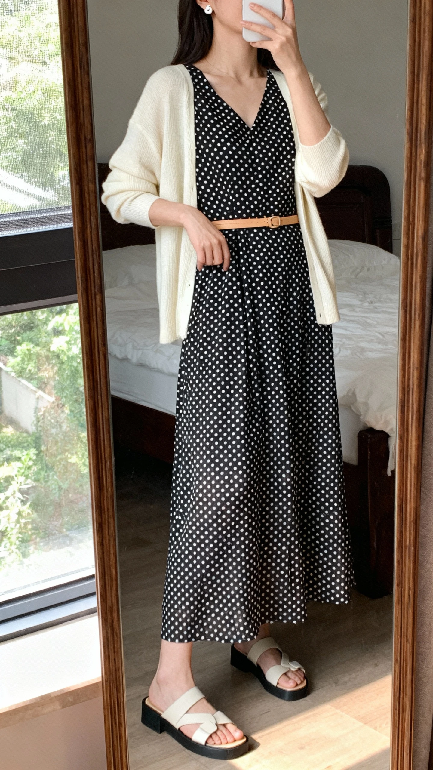 iPhone mirror selfie of a woman wearing a black-and-white polka dot midi-maxi dress with a slim tan belt, soft cream cardigan layered over, simple stud earrings, and low block sandals, face not visible, cropped at neck, bedroom mirror, natural window light, iPhone photo quality.