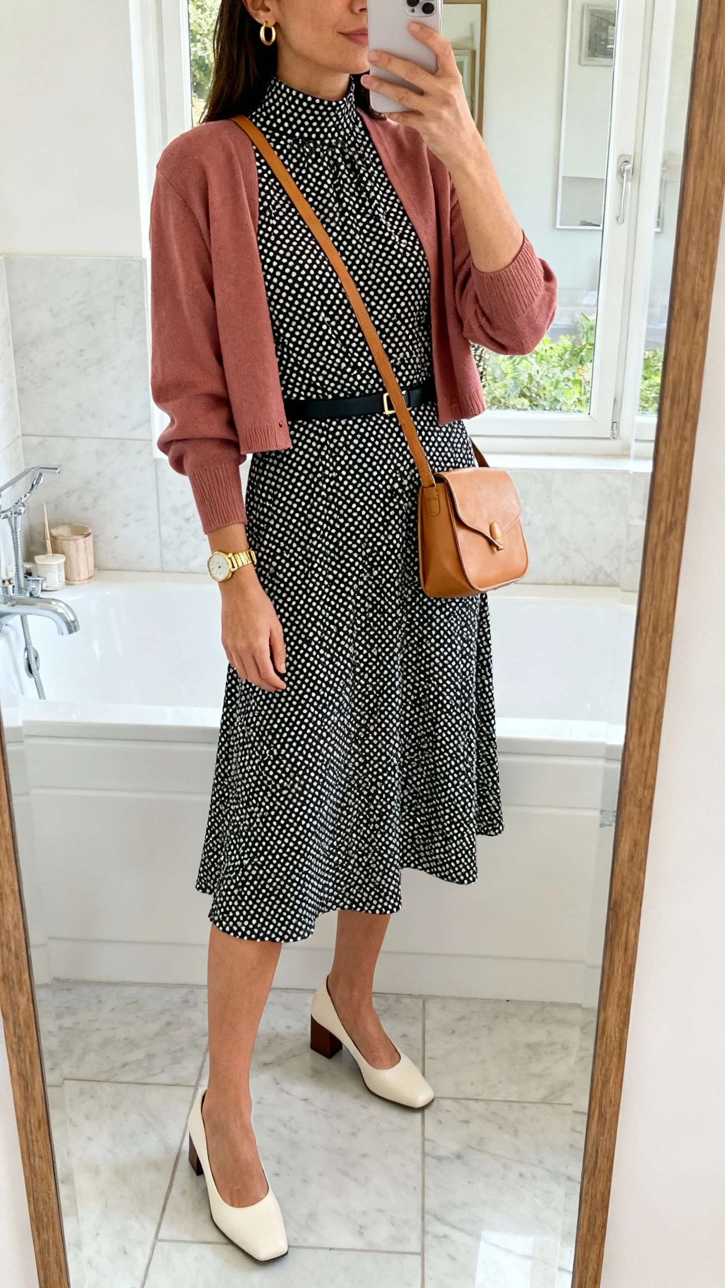 iPhone mirror selfie of a woman wearing a black-and-white polka dot A-line dress with a high neckline, a cropped cardigan in dusty rose, low block-heel slingbacks in cream, a tan leather crossbody bag, small gold hoop earrings, a simple gold watch, and a thin black waist belt, face not visible, bathroom full-length mirror, indoor natural light, iPhone photo quality.