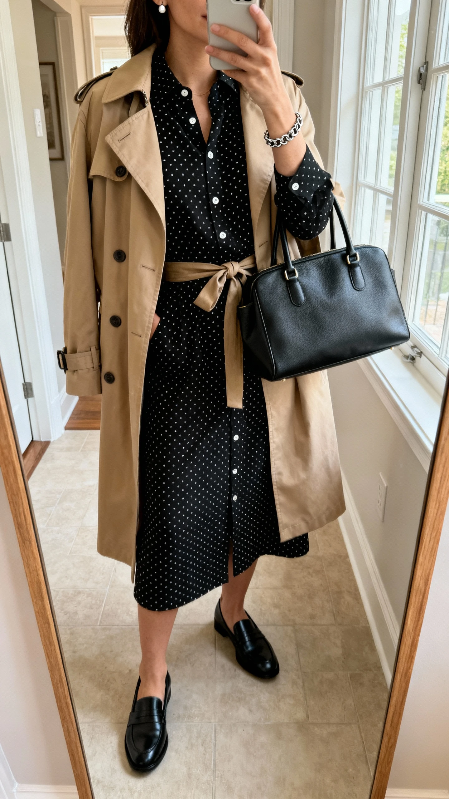iPhone mirror selfie of a woman wearing a black polka dot shirt dress (button-front, midi length), black leather loafers, a beige classic trench coat, a medium black satchel, simple stud earrings, a chain bracelet, and a tied waist belt, face not visible, cropped at neck, entryway full-length mirror, indoor window light, iPhone photo quality.