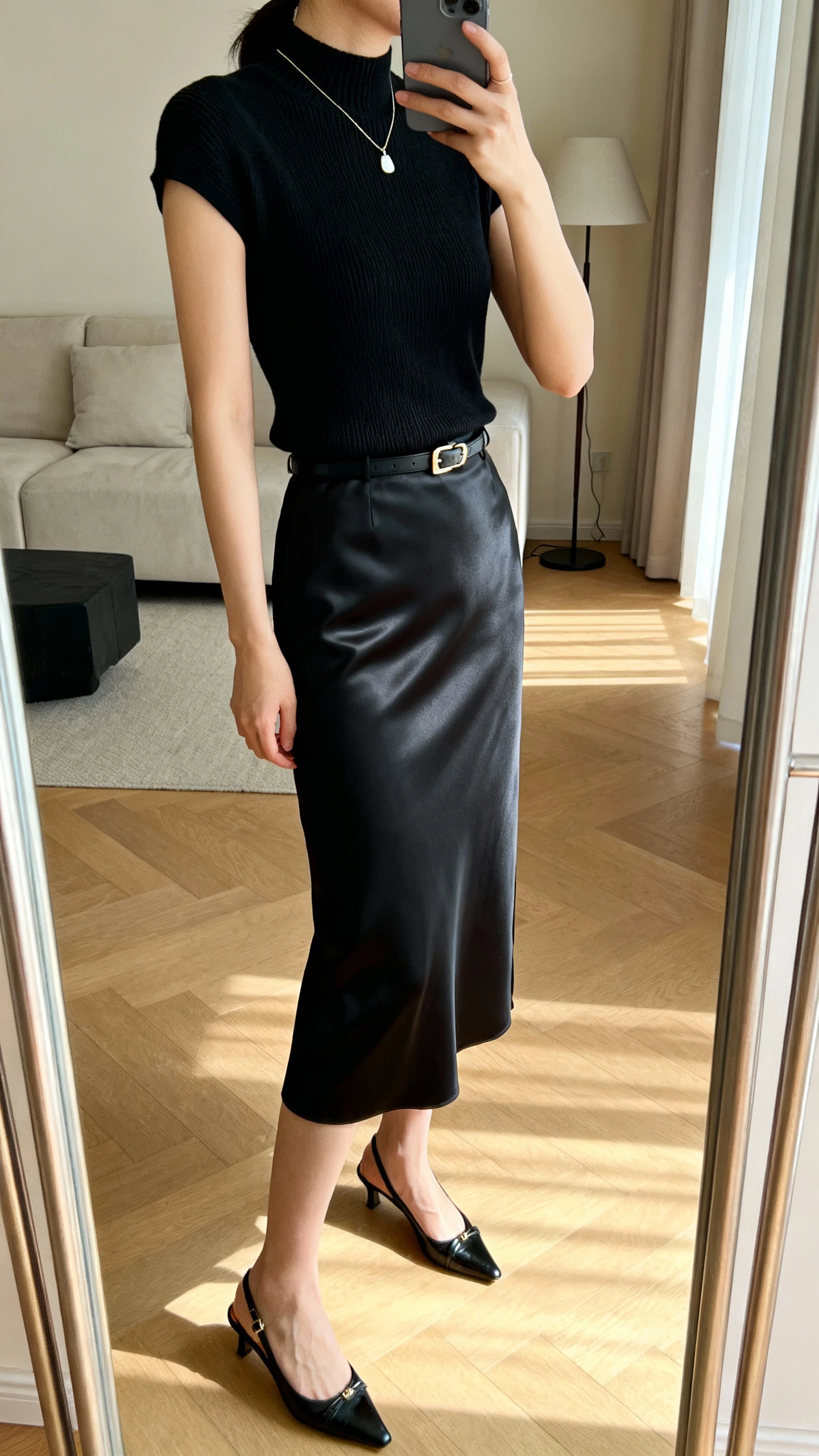 iPhone mirror selfie of a woman wearing a black satin column skirt, a short-sleeve structured knit tee (mock neck), elegant slingbacks, a refined belt under the tee, and a simple pendant necklace, face not visible, side angle, full-length mirror in living room, soft morning light, iPhone photo quality.