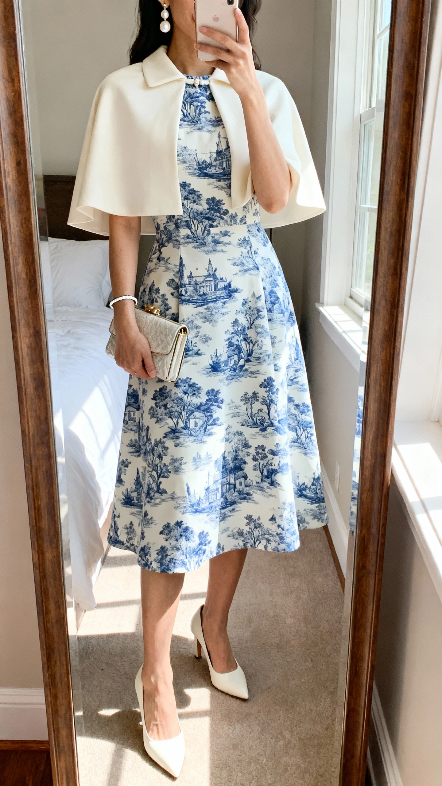 iPhone mirror selfie of a woman wearing a blue-and-white toile-print A-line midi dress with a modest neckline, cream capelet, cream kitten-heel pumps, elegant pearl drop earrings, slim bracelet, and a structured clutch, face not visible, cropped at shoulders, bedroom mirror with natural window light, iPhone photo quality.