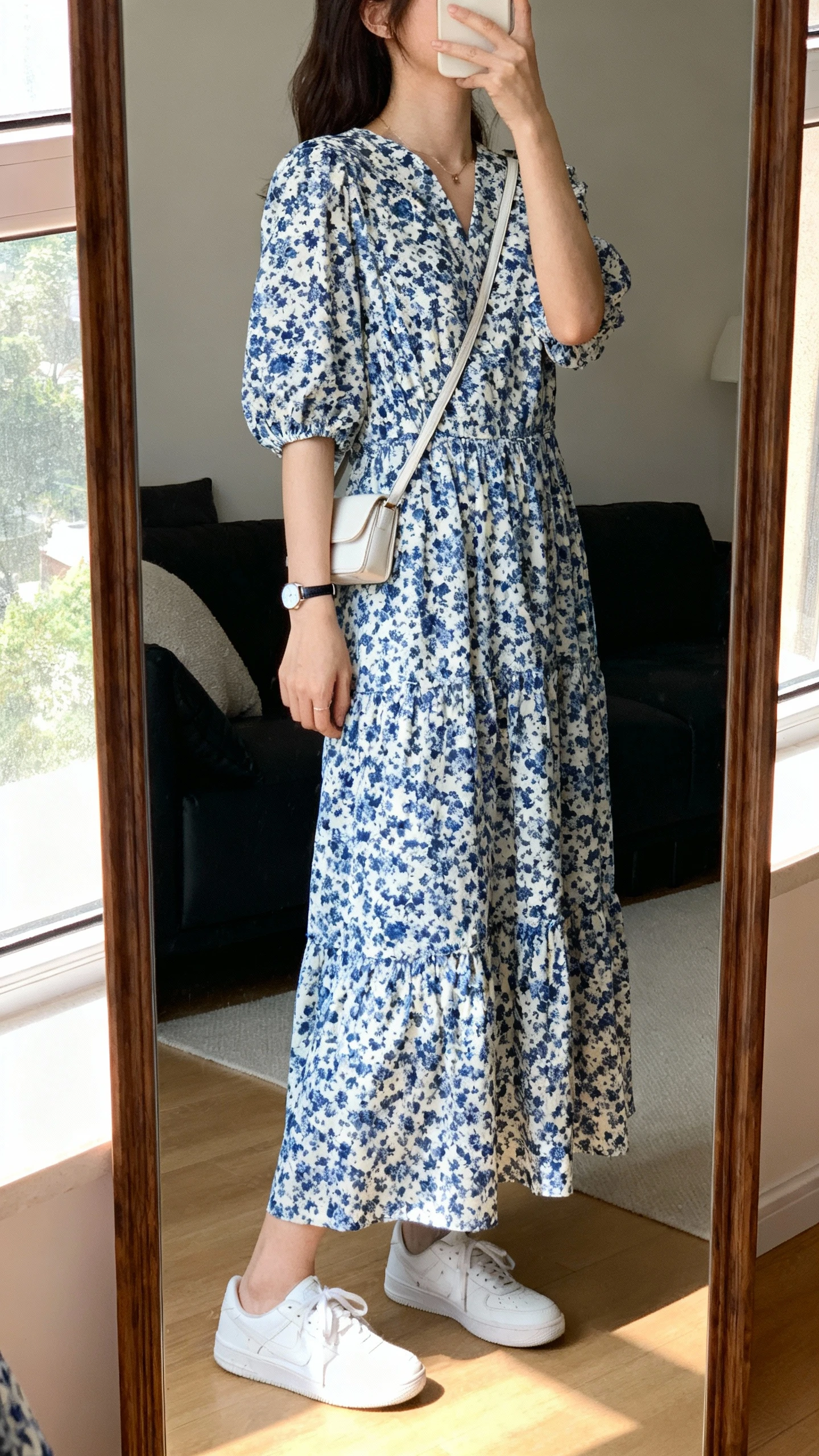 iPhone mirror selfie of a woman wearing a blue-and-white floral maxi dress with clean white sneakers, small crossbody and simple watch, face not visible, side angle, living room full-length mirror, soft afternoon window light, iPhone photo quality.