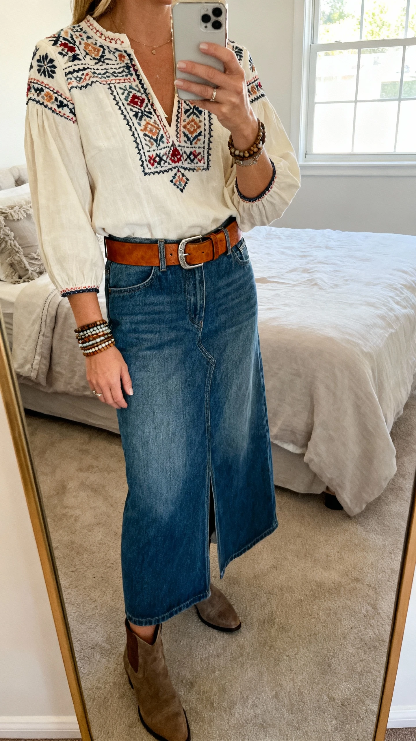 iPhone mirror selfie of a woman wearing a boho embroidered blouse tucked into a denim maxi skirt with a wide leather belt, stacked bracelets, and ankle boots, face not visible, cropped at neck, full-length mirror in a cozy bedroom, natural window light, iPhone photo quality.