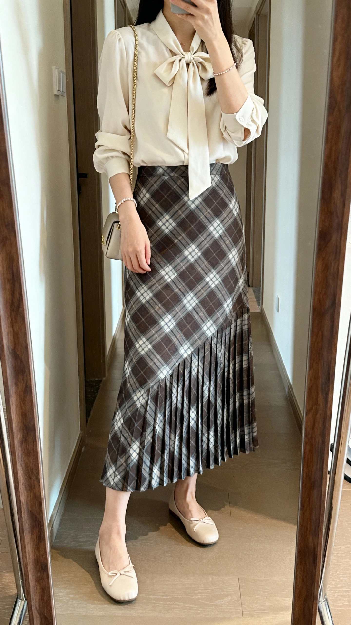 iPhone mirror selfie of a woman wearing a bow-tie blouse paired with a pleated plaid maxi skirt, ballet flats, dainty bracelet, and a small shoulder bag, face not visible, cropped at shoulders, full-length mirror in hallway, natural daylight, iPhone photo quality.