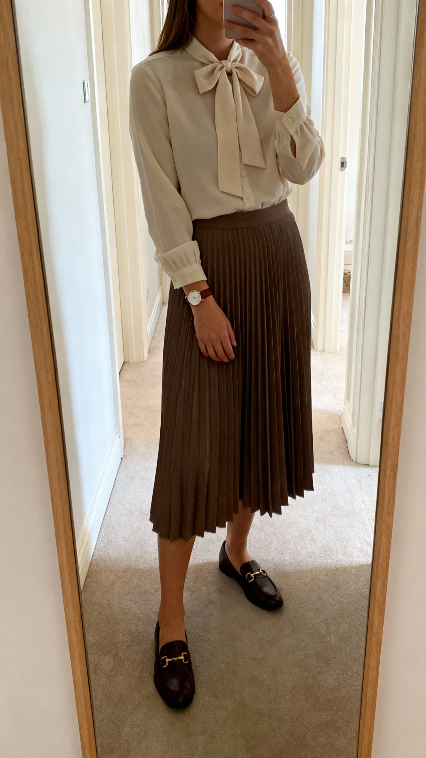 iPhone mirror selfie of a woman wearing a bow-tie neck blouse with long sleeves and a pleated midi skirt, classic loafers and a slim leather watch, face not visible, cropped at shoulders, full-length hallway mirror, soft daylight, iPhone photo quality.