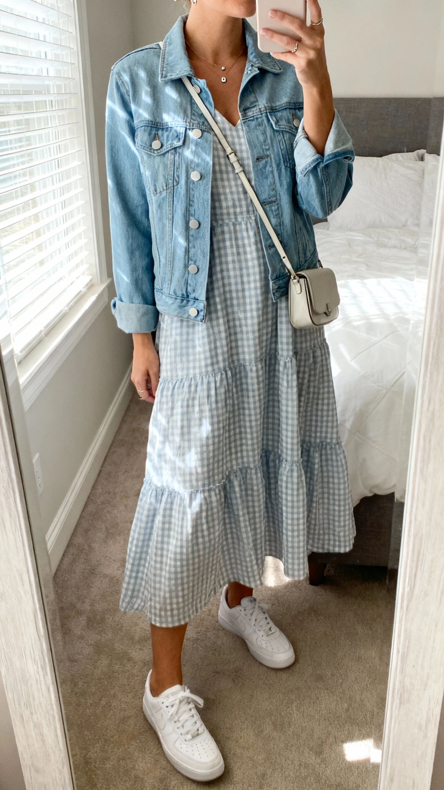 iPhone mirror selfie of a woman wearing a breezy gingham check dress with a light blue denim jacket, simple white sneakers, small crossbody bag, and minimal studs, face not visible, cropped at neck, bedroom mirror, soft morning window light, iPhone photo quality.