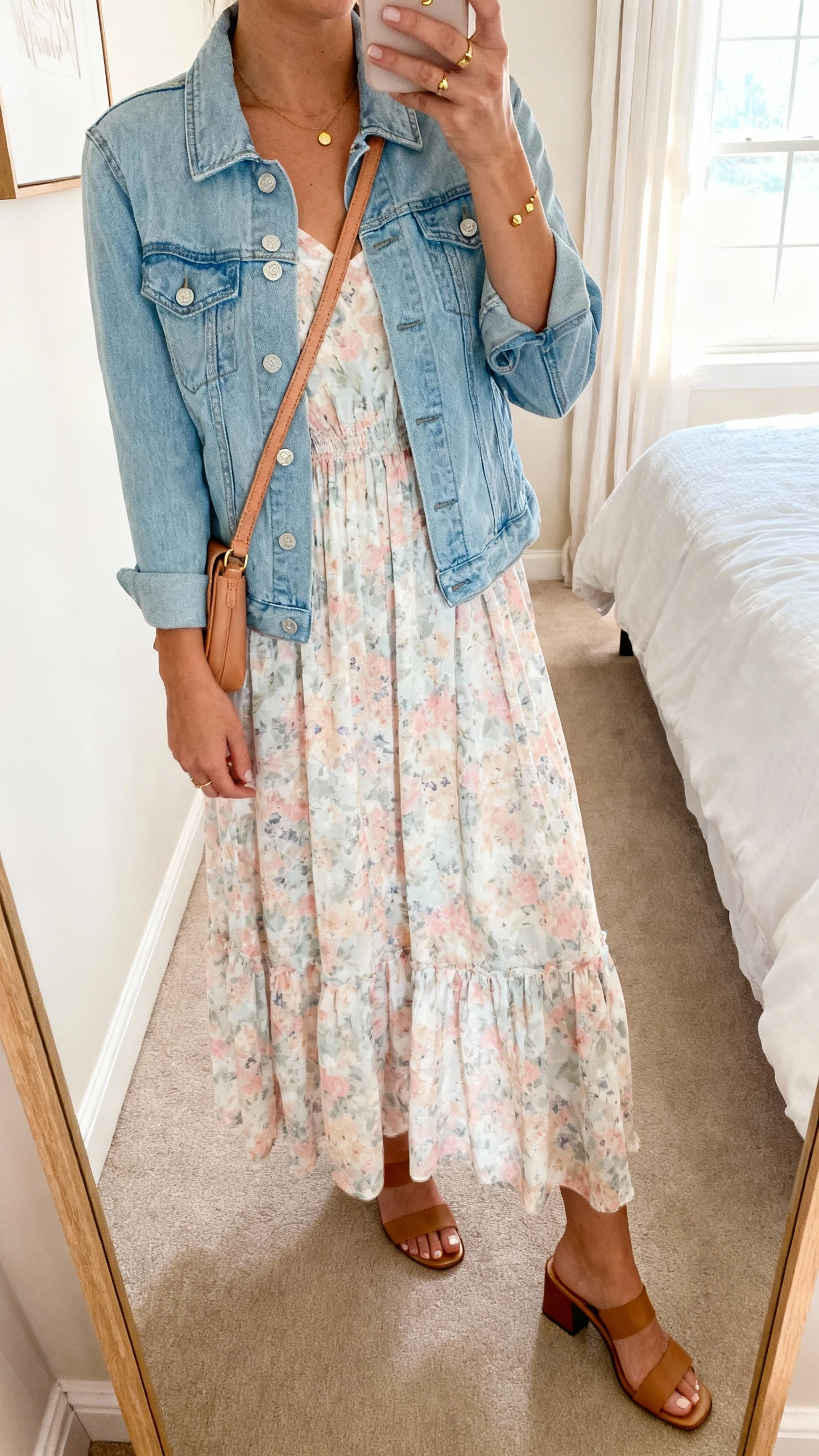 iPhone mirror selfie of a woman wearing a breezy pastel floral maxi dress with a light blue denim jacket, simple gold studs, a tan crossbody, and low block-heel sandals, face not visible, cropped at neck, bedroom mirror, soft morning window light, iPhone photo quality.