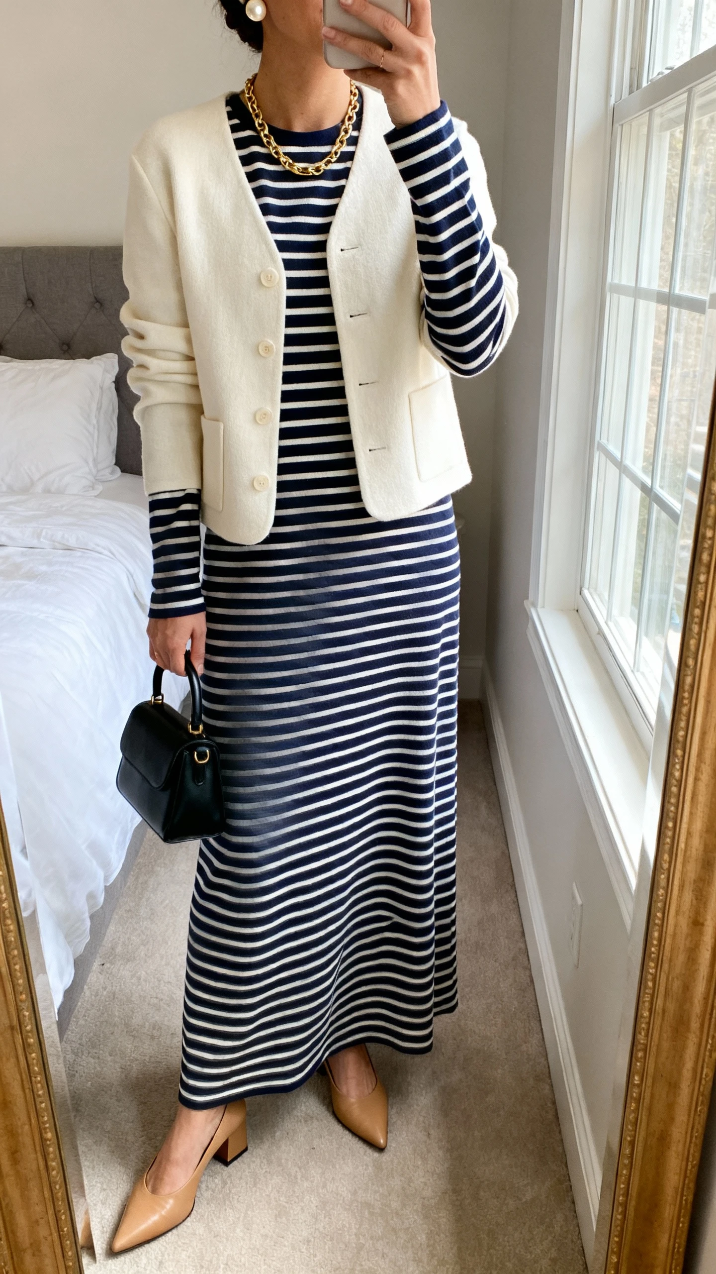 iPhone mirror selfie of a woman wearing a Breton-striped long-sleeve maxi dress in cotton (navy/white), structured cream cardigan, pointed-toe block-heel pumps in tan, delicate pearl stud earrings, simple gold chain necklace, and a small black top-handle bag; face not visible, cropped at neck, bedroom mirror, natural window light, iPhone photo quality.