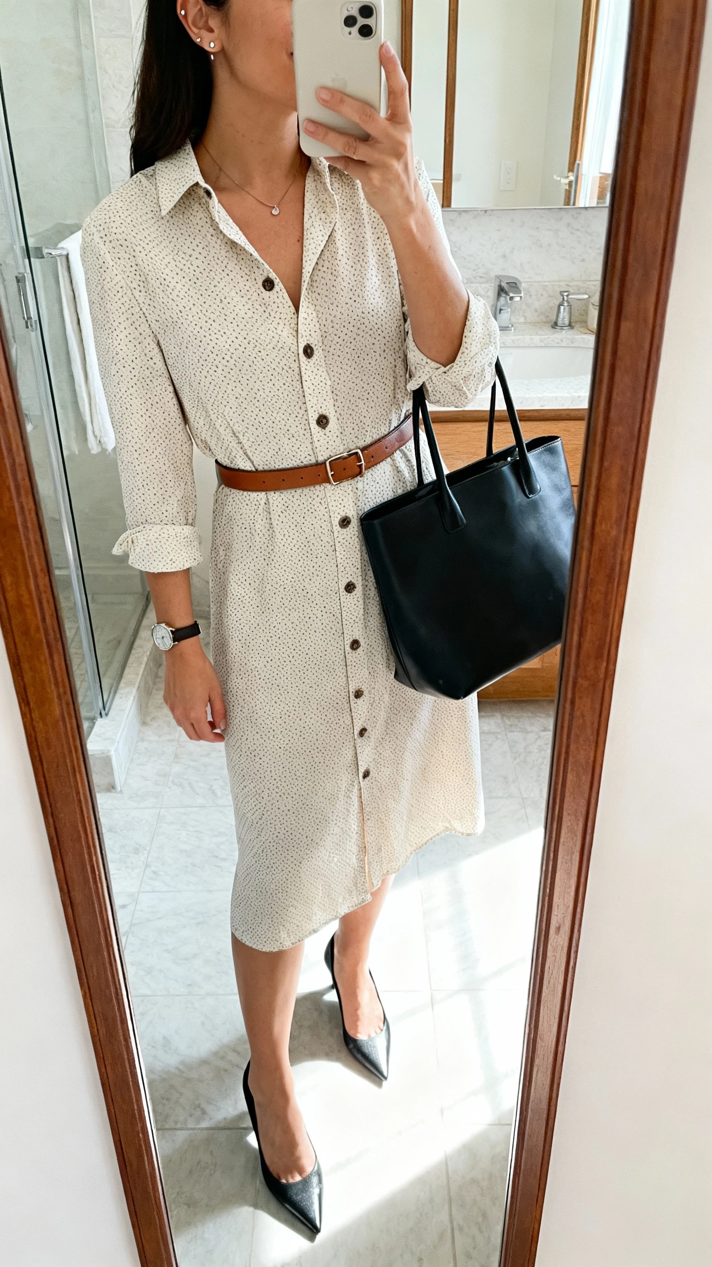 iPhone mirror selfie of a woman wearing a button-front shirt dress in a micro-dot print, a slim leather belt matched to pointed-toe kitten heels, minimal stud earrings, a thin watch, a subtle pendant, and a structured black tote bag; face not visible, cropped at shoulders, bathroom mirror, morning light, iPhone photo quality.