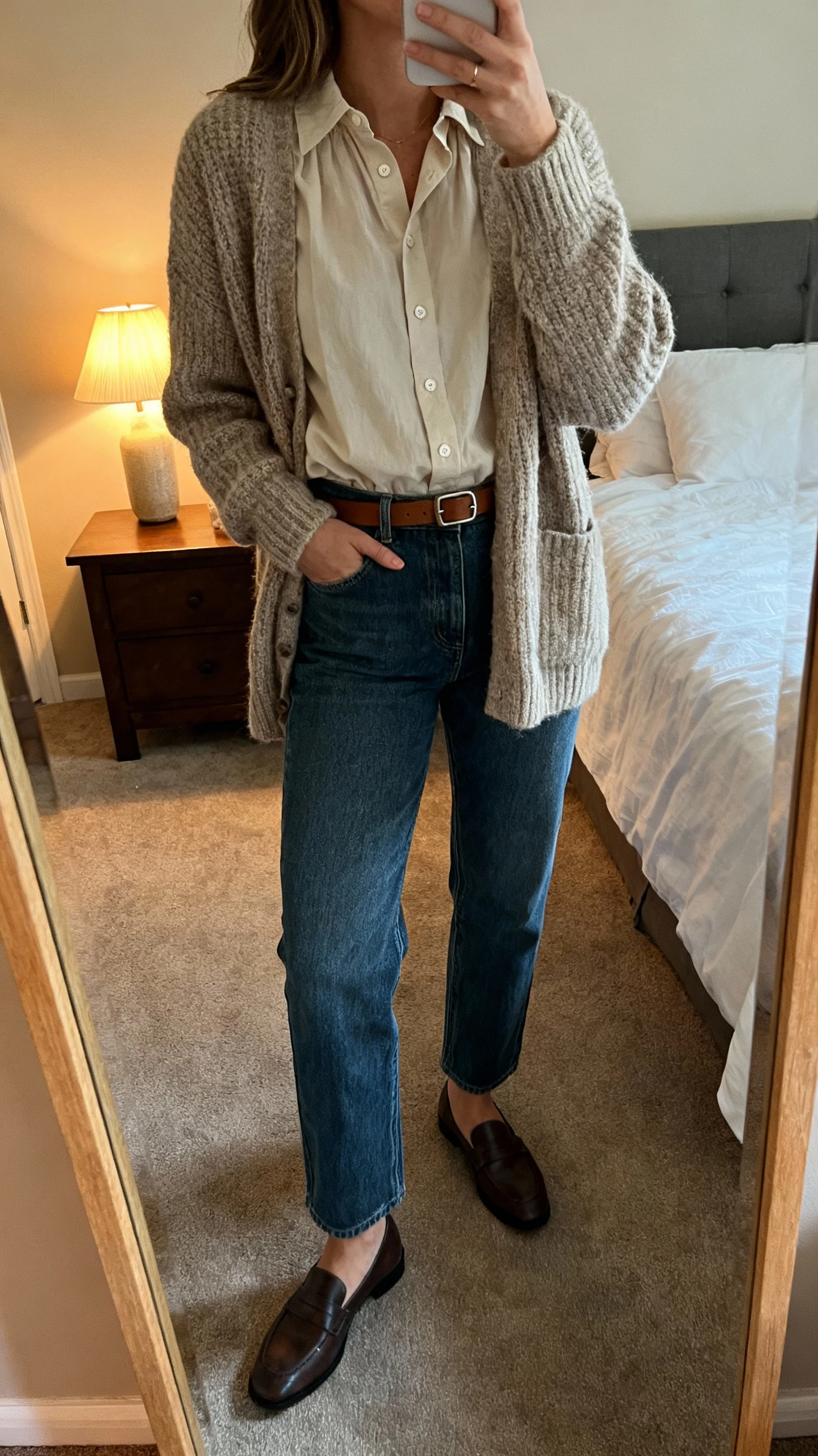 iPhone mirror selfie of a woman wearing a buttoned-up poplin blouse under a cozy knit cardigan, straight-leg jeans, loafers, and a minimal leather belt, face not visible, cropped at shoulders, bedroom mirror, warm indoor lamp light, iPhone photo quality.
