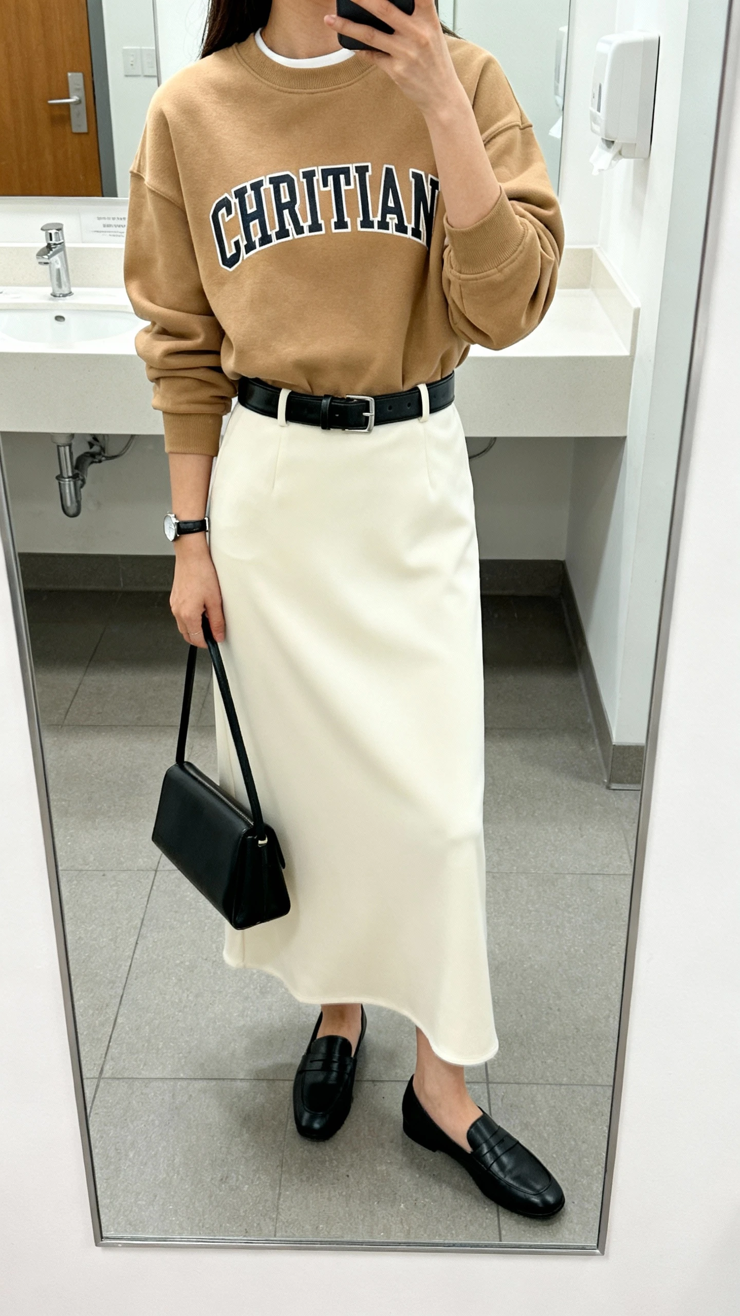 iPhone mirror selfie of a woman wearing a camel Christian crewneck over a sleek cream maxi dress, black leather belt to define waist, minimalist black loafers, slim black tote, and a simple watch, face not visible, cropped at neck, office bathroom mirror, bright indoor lighting, iPhone photo quality