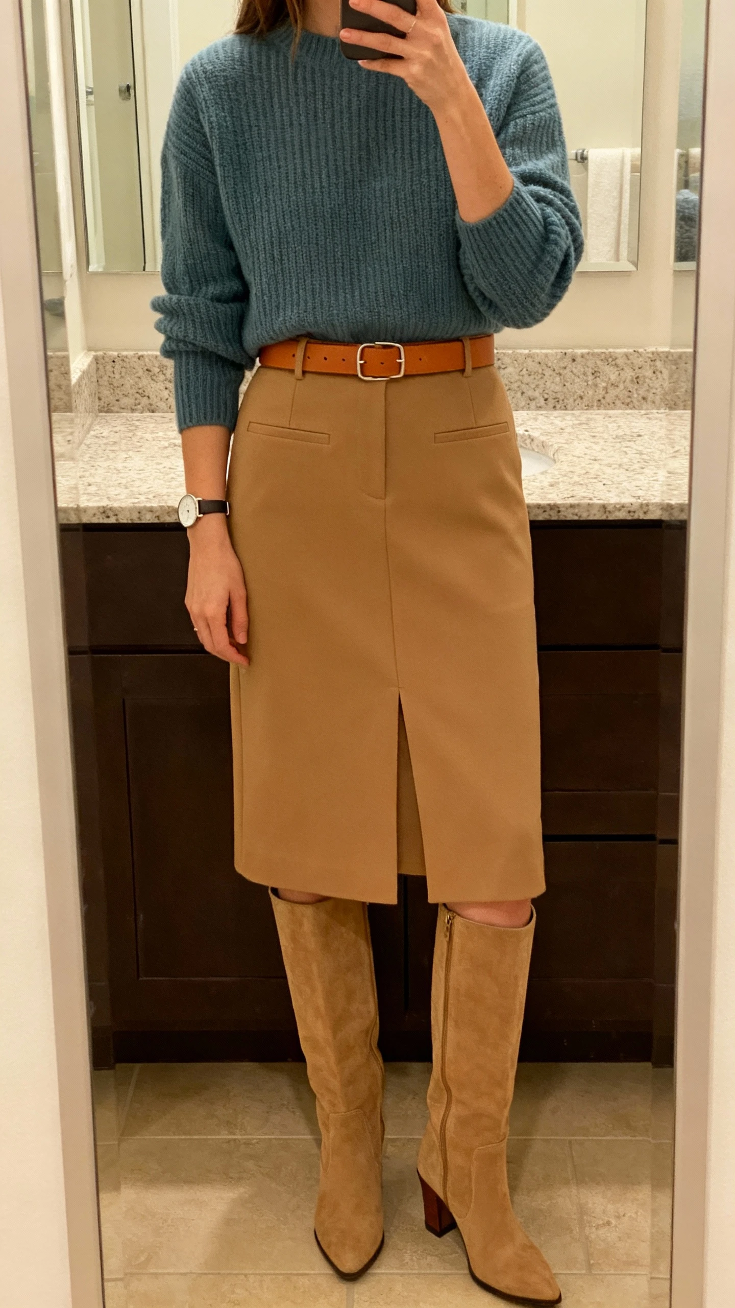 iPhone mirror selfie of a woman wearing a camel structured midi skirt, a fine-knit dusty blue sweater, knee-high low-heel boots, a matching camel leather belt, and a minimalist watch, face not visible, cropped at shoulders, bathroom mirror, indoor warm lighting, iPhone photo quality.