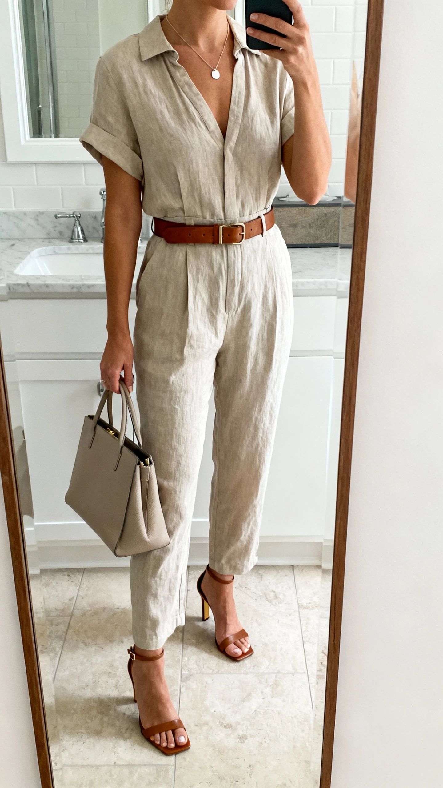 iPhone mirror selfie of a woman wearing a casual-cool cotton jumpsuit cinched with a leather belt, elevated with heeled sandals, a minimal pendant necklace, and a refined tote, face not visible, cropped at neck, bathroom mirror, natural daylight, iPhone photo quality.