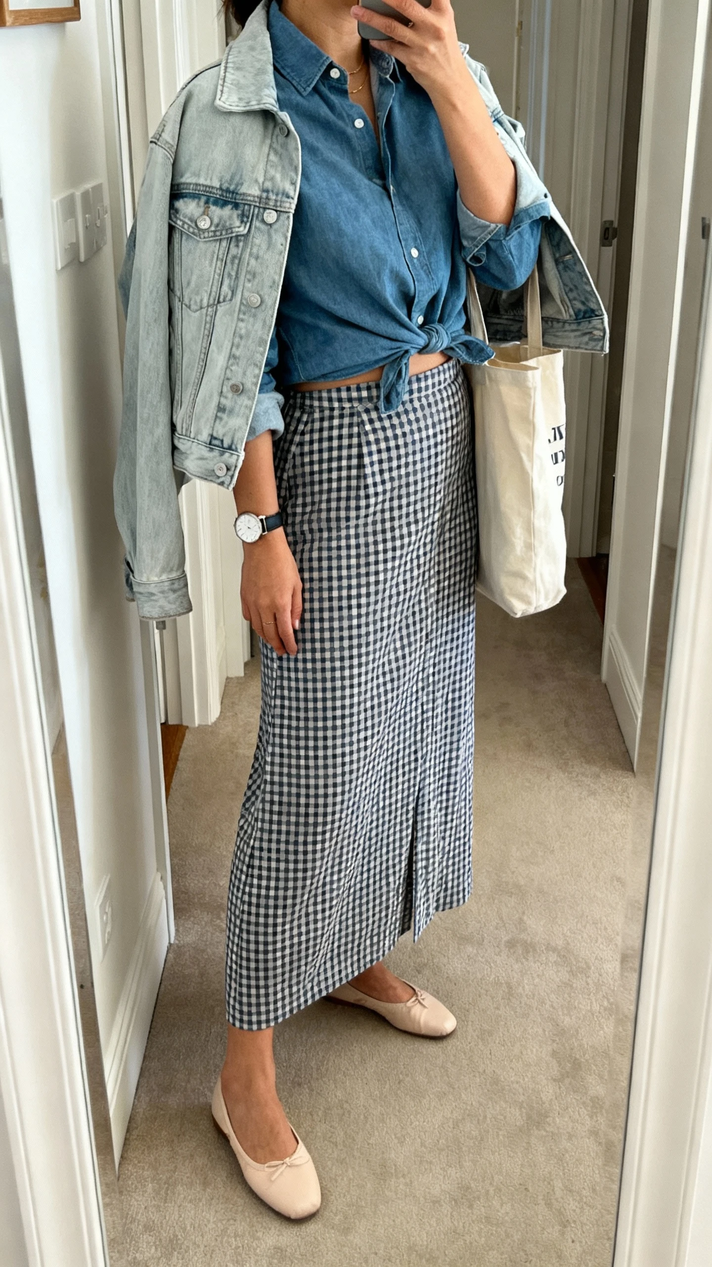 iPhone mirror selfie of a woman wearing a chambray shirt half-tucked into a gingham long skirt, light-wash denim jacket over shoulders, comfortable ballet flats, simple wristwatch, and a tote bag; face not visible, side angle, entryway mirror, soft indoor lighting, iPhone photo quality.