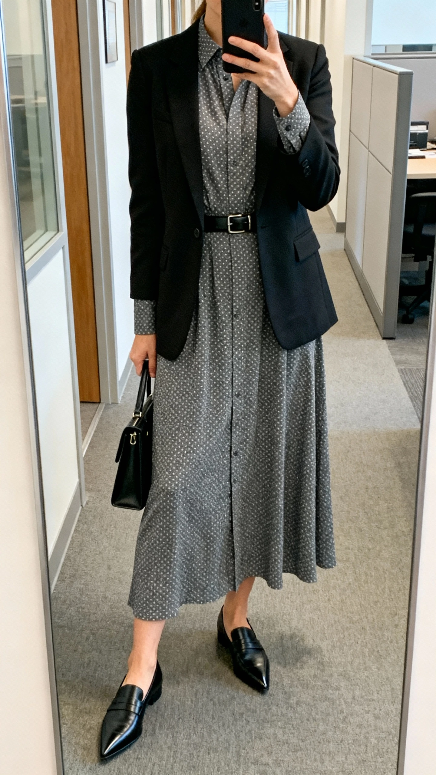iPhone mirror selfie of a woman wearing a charcoal polka dot long-sleeve shirt dress with calf-grazing hem, tailored black blazer, pointed-toe black loafers, slim black leather belt at the waist, and a slim black work tote, face not visible, cropped at shoulders, office hallway mirror, indoor lighting, iPhone photo quality.
