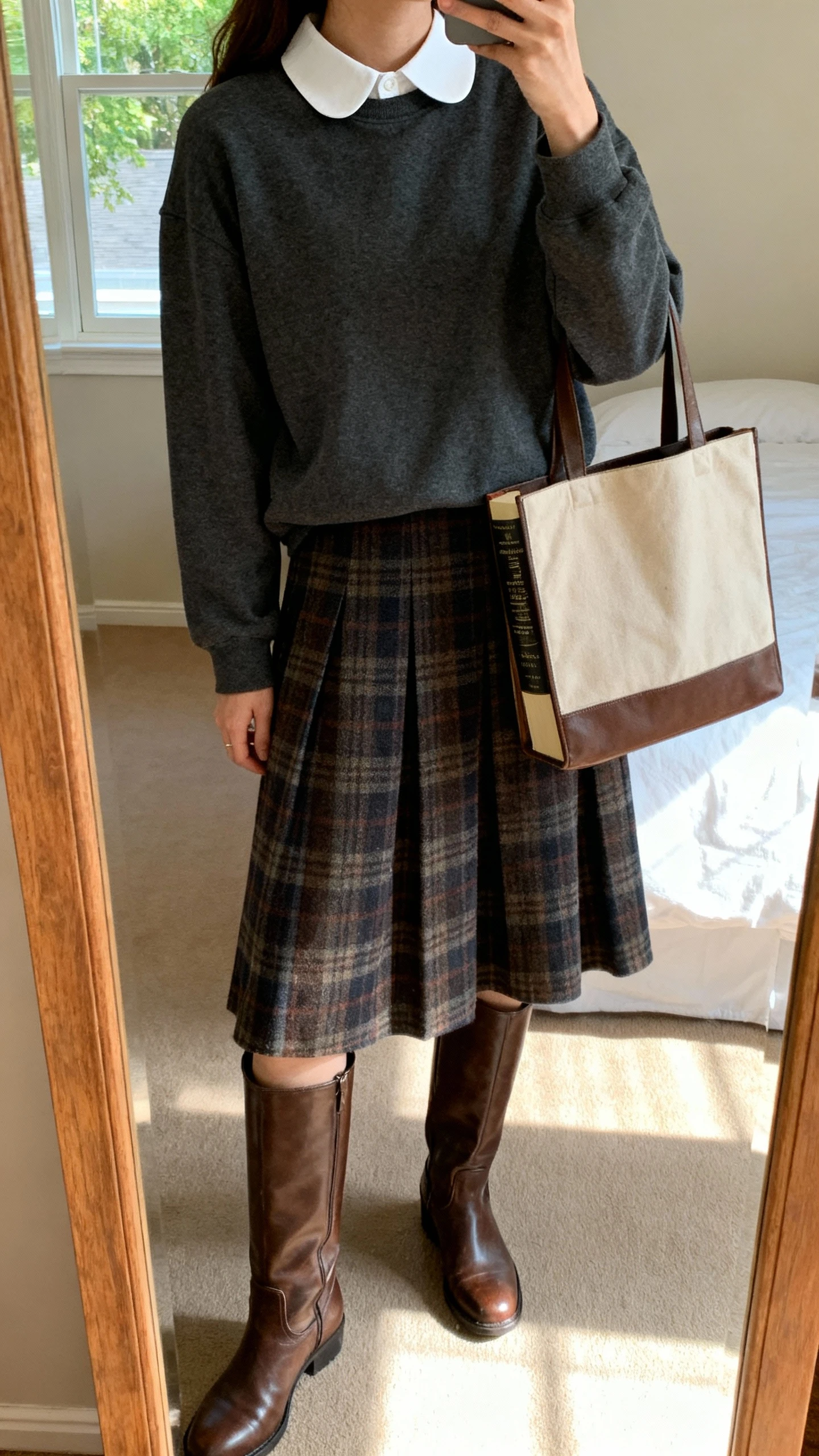 iPhone mirror selfie of a woman wearing a charcoal crewneck layered over a white Peter Pan–collar blouse, plaid A-line midi skirt, knee-high leather boots, and a bookish tote, face not visible, cropped at shoulders, bedroom mirror, afternoon window light, iPhone photo quality.
