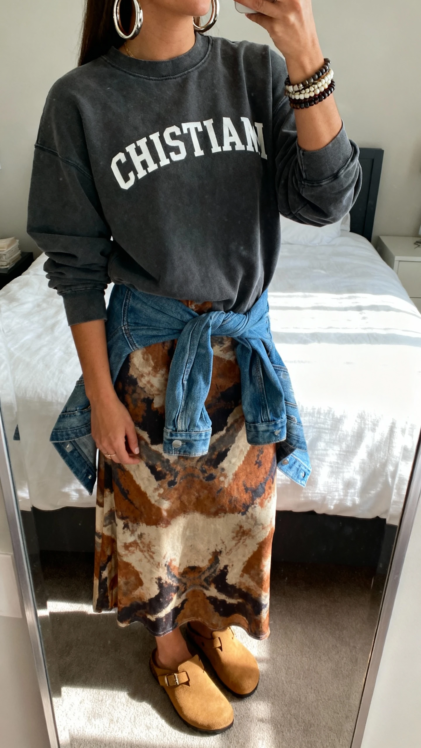 iPhone mirror selfie of a woman wearing a charcoal Christian crewneck over a patterned artsy maxi dress (abstract earthy tones), chunky hoop earrings, stack of bracelets, denim jacket tied at waist, and tan clogs, face not visible, cropped at shoulders, bedroom mirror, diffused daylight, iPhone photo quality