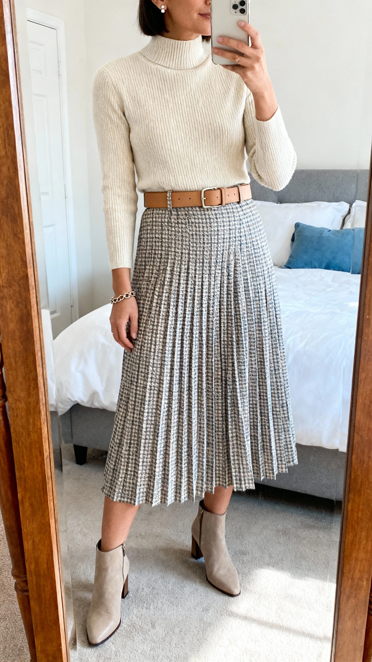 iPhone mirror selfie of a woman wearing a checkered A-line or pleated midi skirt in soft tones (stone/cream, taupe/ivory, or misty blue), a fine-gauge knit top or mock-neck sweater in cream or sand, optional classic camel belt, heeled ankle boots or sleek loafers, minimalist stud earrings, and a delicate chain bracelet; face not visible, cropped at shoulders, bedroom mirror, warm indoor lighting, iPhone photo quality.