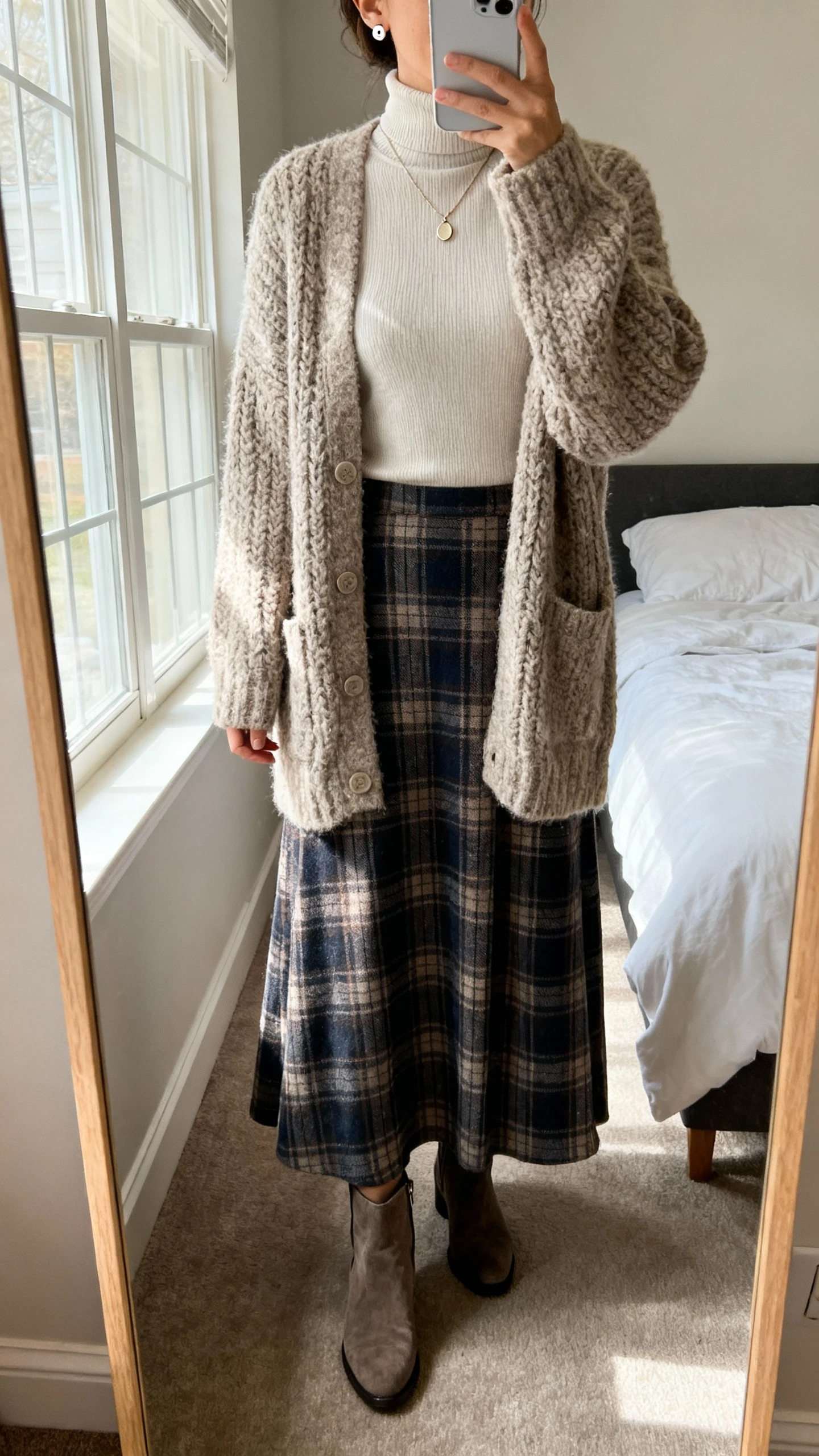 iPhone mirror selfie of a woman wearing a classic plaid long skirt with a chunky cozy cardigan layered over a fitted turtleneck, simple stud earrings, delicate pendant necklace, and ankle boots, face not visible, cropped at neck, bedroom mirror, morning window light, iPhone photo quality.
